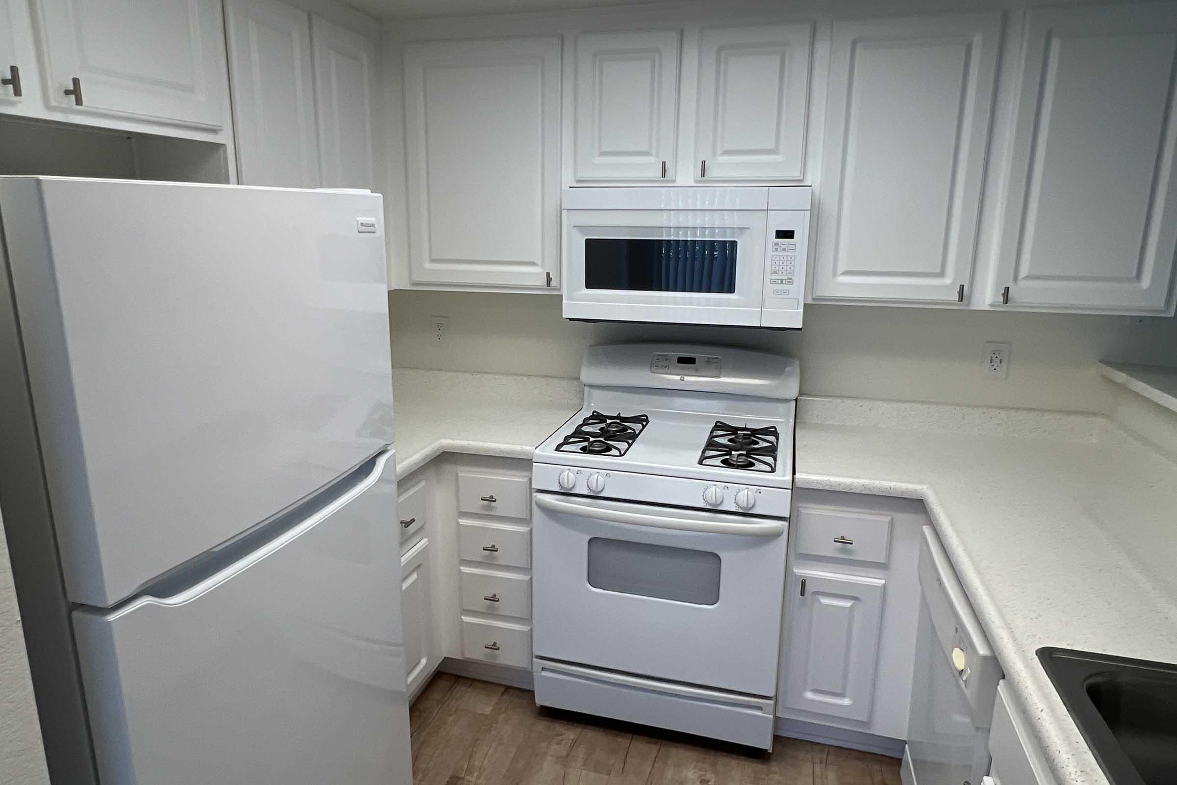 A modern kitchen featuring white cabinetry, a white refrigerator, a built-in microwave above a gas stove, and a light-colored countertop. The layout is compact with ample storage space and a clean, bright aesthetic. The flooring is a light wood finish, complementing the overall design.