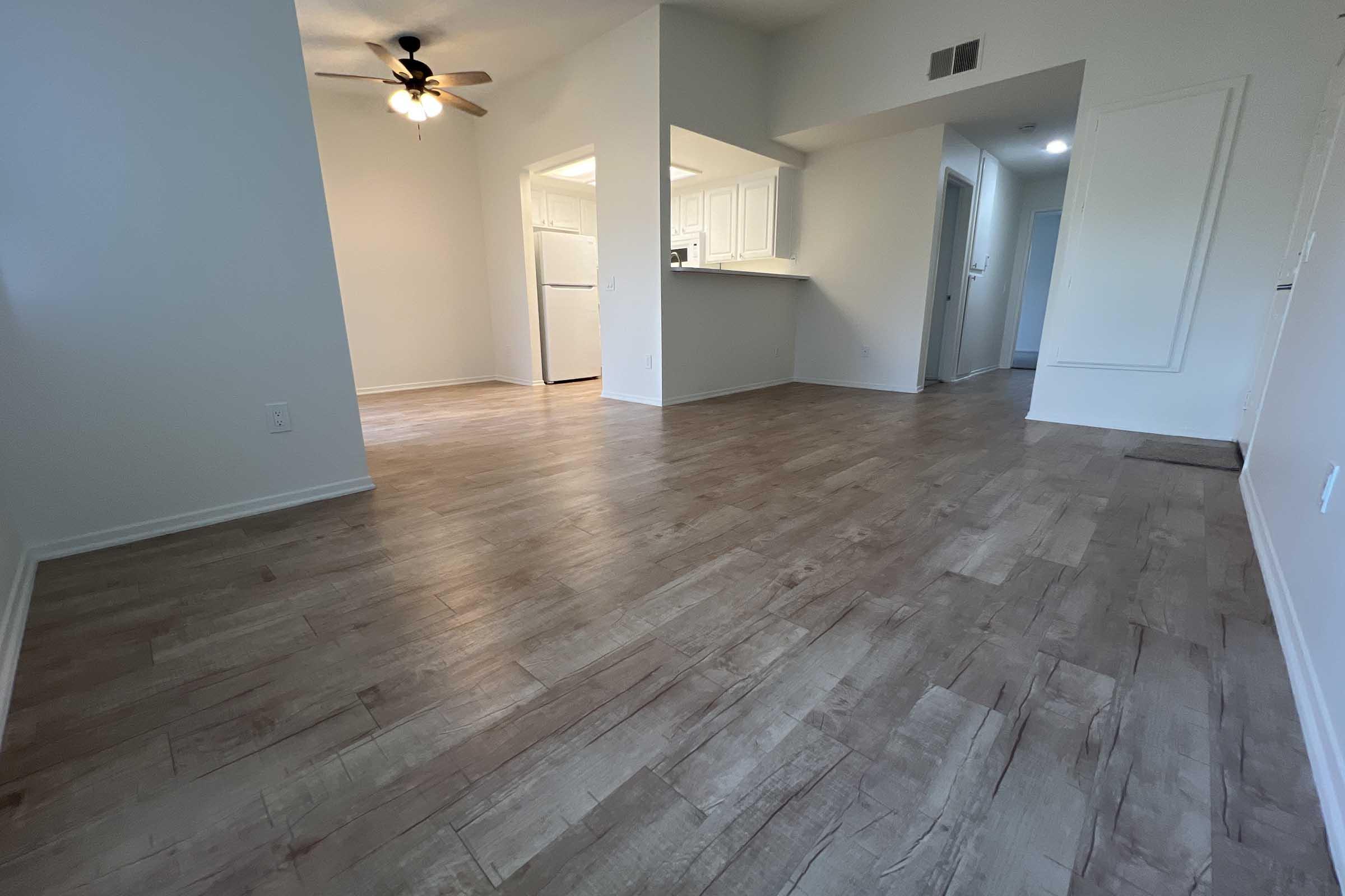 Interior view of a spacious, well-lit living area featuring a light-colored laminate floor, a ceiling fan, and an open layout leading to a kitchen with white cabinetry. The walls are painted in a neutral tone, enhancing the airy feel of the space.