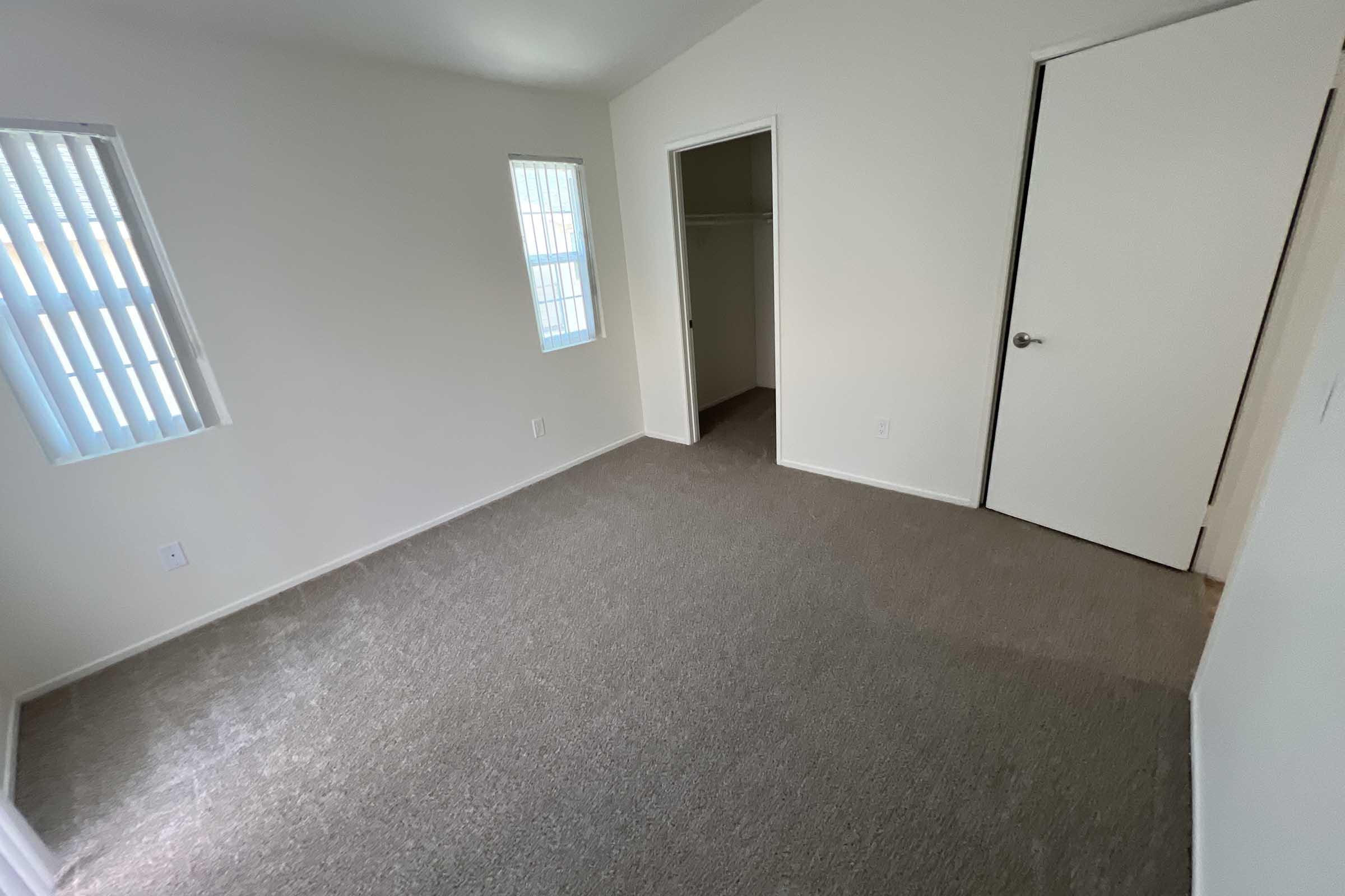 A simple, empty room with beige carpet and light-colored walls. It features two small windows on the left side and a closet with a door on the right. The room has a sloped ceiling, creating a spacious feel despite its minimal furnishings. Natural light enters through the windows, enhancing the bright atmosphere.