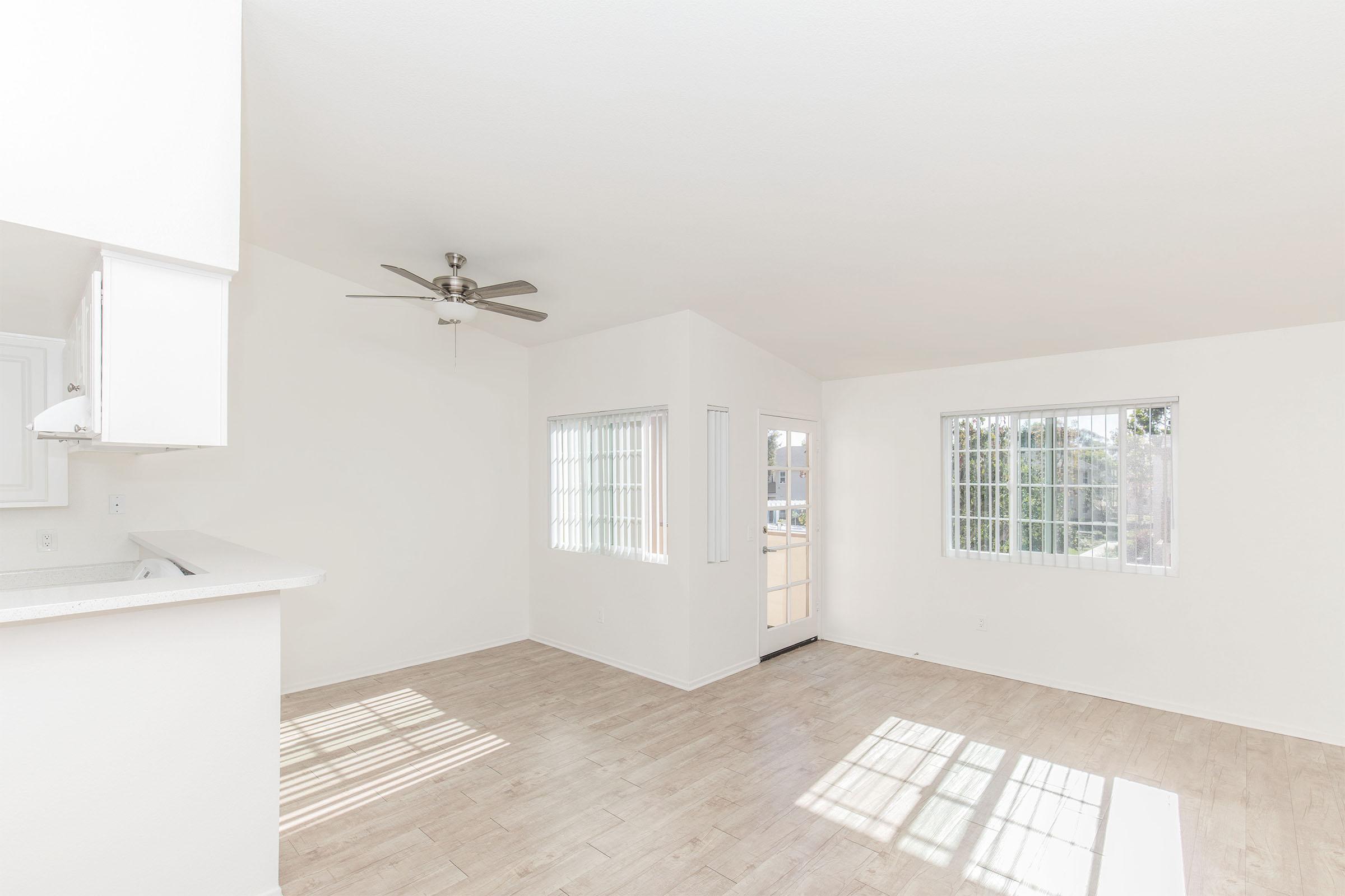 A spacious, bright living area featuring light-colored walls and light wood flooring. Two large windows allow natural light to fill the space, and there's a ceiling fan for ventilation. A doorway leads to an exterior entry, and the adjacent kitchen area is partially visible.
