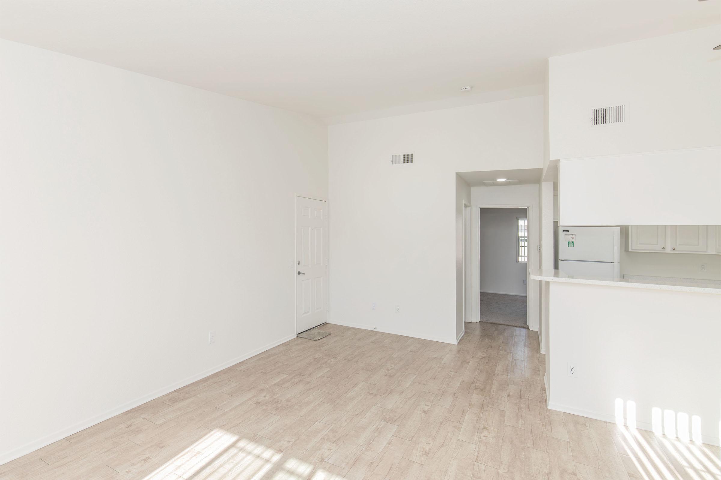 A spacious and bright living area with light-colored walls and wooden flooring. There's a doorway leading to another room, a kitchenette with white cabinetry visible in the background, and windows allowing natural light to enter the space. The room appears clean and uncluttered, suitable for a modern apartment.