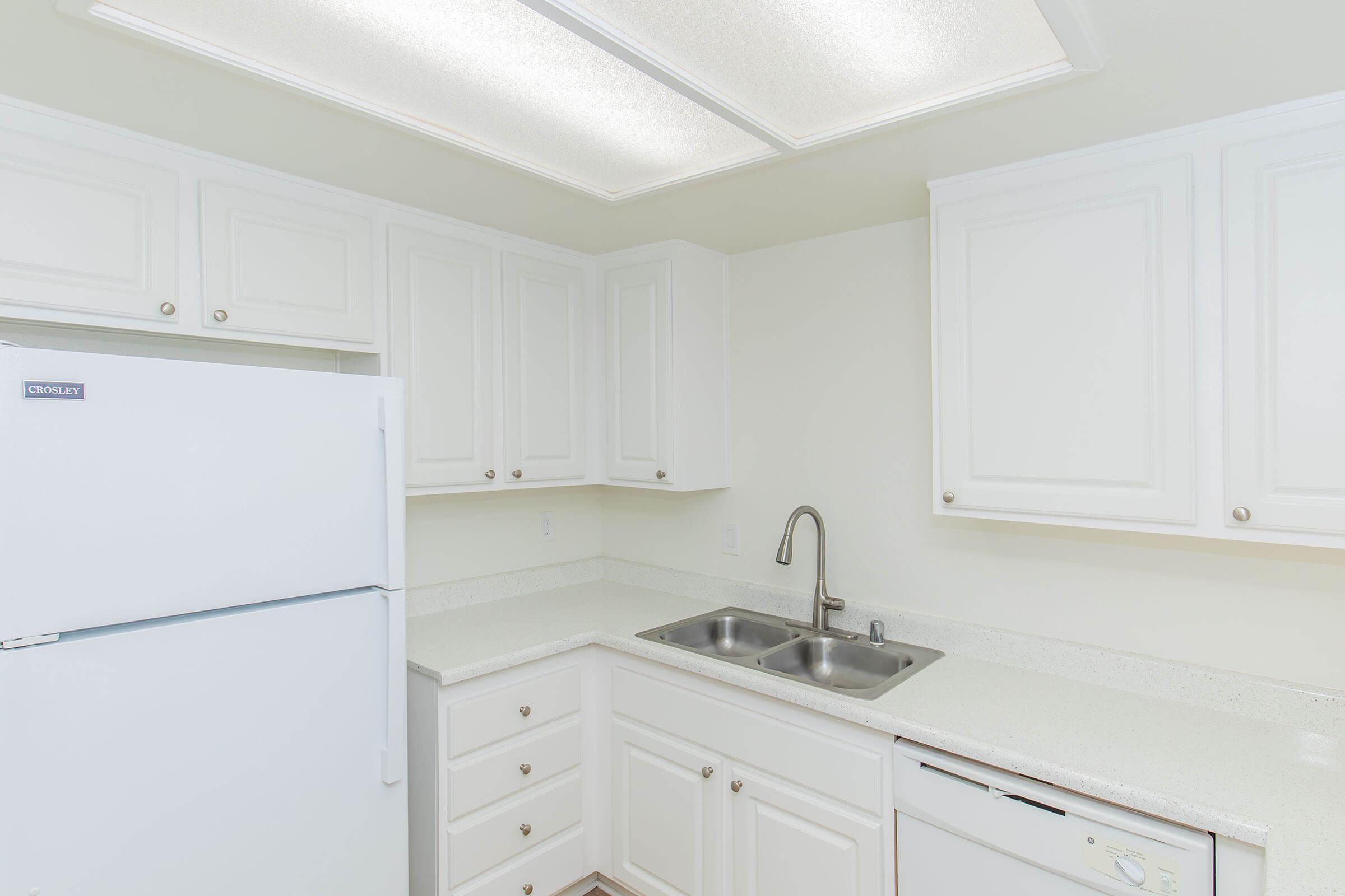A clean, modern kitchen with white cabinetry, a double sink, and a light-colored countertop. The refrigerator is on the left side, and a built-in dishwasher is next to the sink. The ceiling features a textured panel design, and the walls are painted a neutral tone.