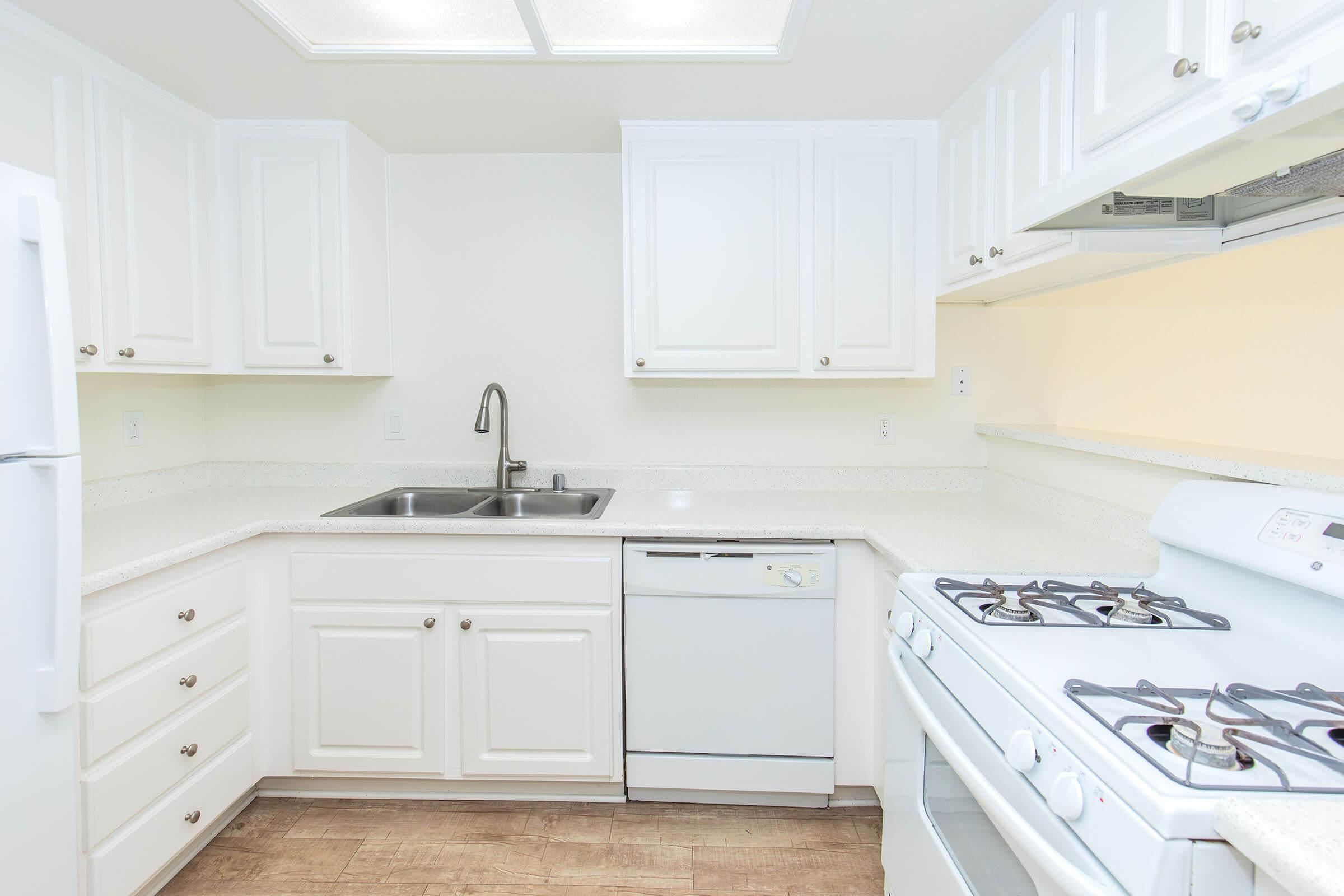 A modern kitchen featuring white cabinetry, a double sink, a dishwasher, and a gas stove. The countertops are light-colored, and the floor has a wood-like finish. The space is bright with ample storage and a clean, minimalist design.