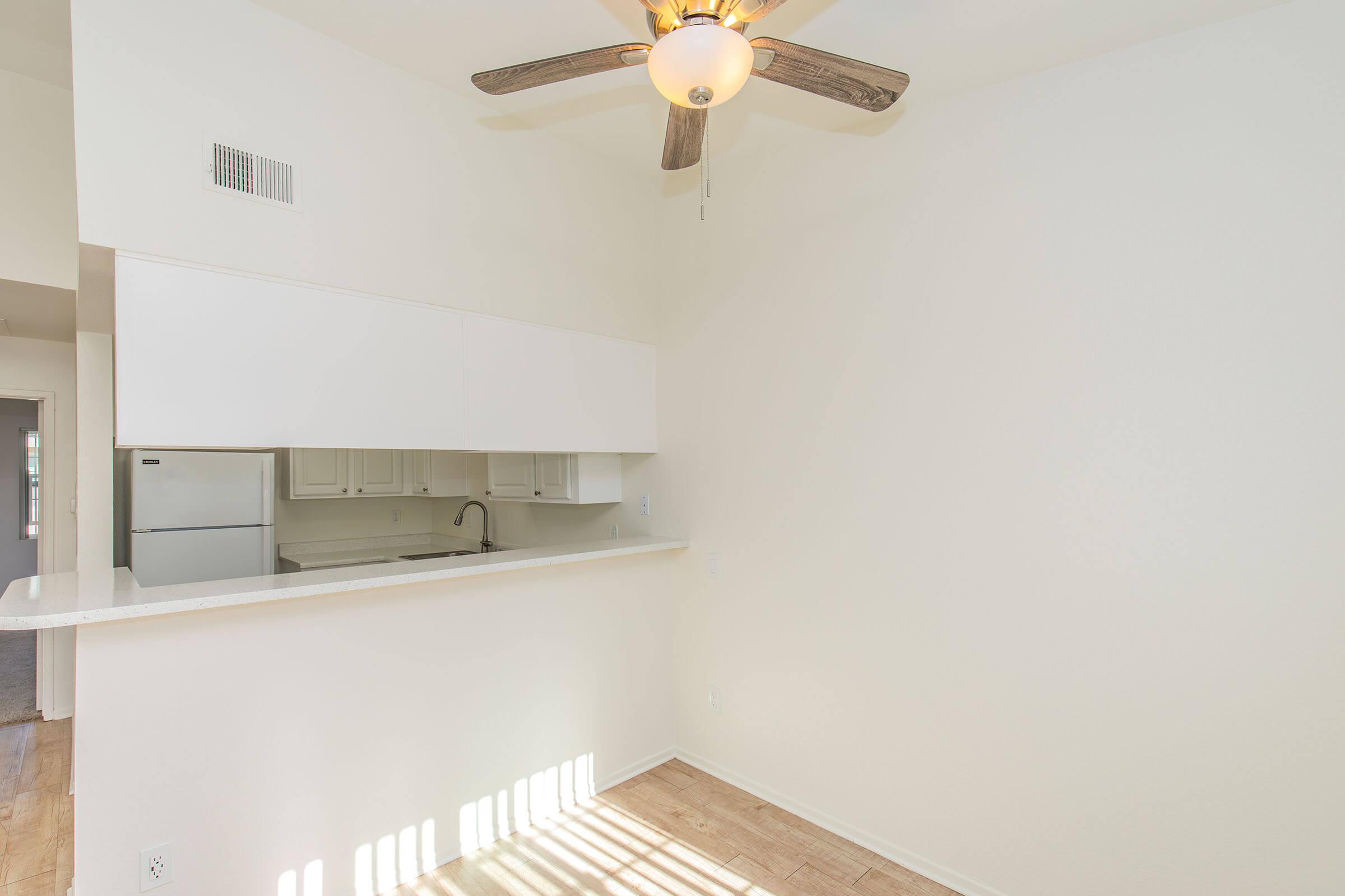 Bright and airy living space featuring a ceiling fan, a bar-height counter separating the kitchen from the main area, and a minimalist design. The walls are painted in light colors, and sunlight streams in, casting shadows on the floor. A refrigerator is visible in the kitchen area.