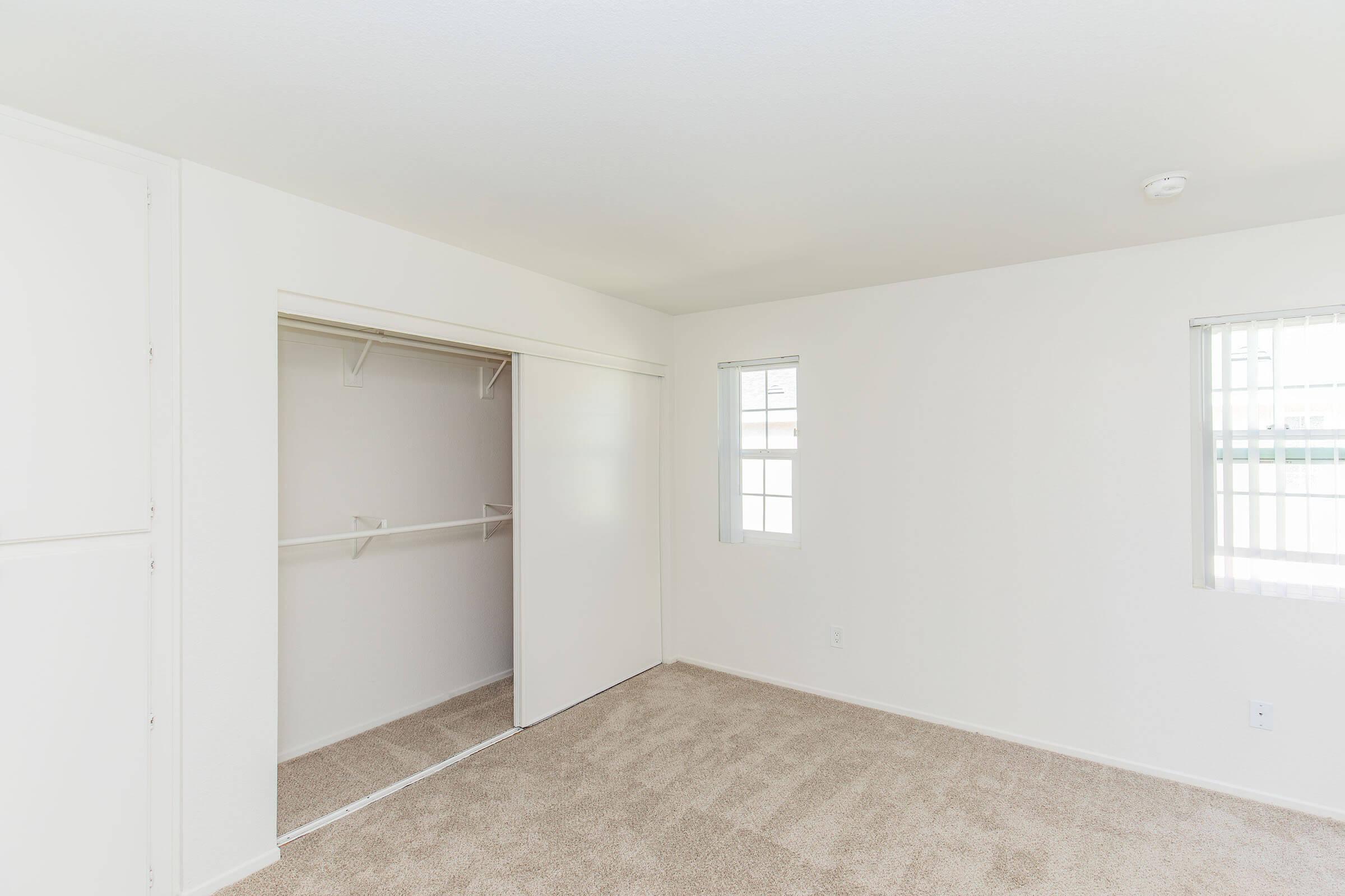 A bright, empty bedroom featuring beige carpeting, two windows with white blinds, and a built-in closet with sliding doors on one side. The walls are painted white, creating a clean and minimalist aesthetic.