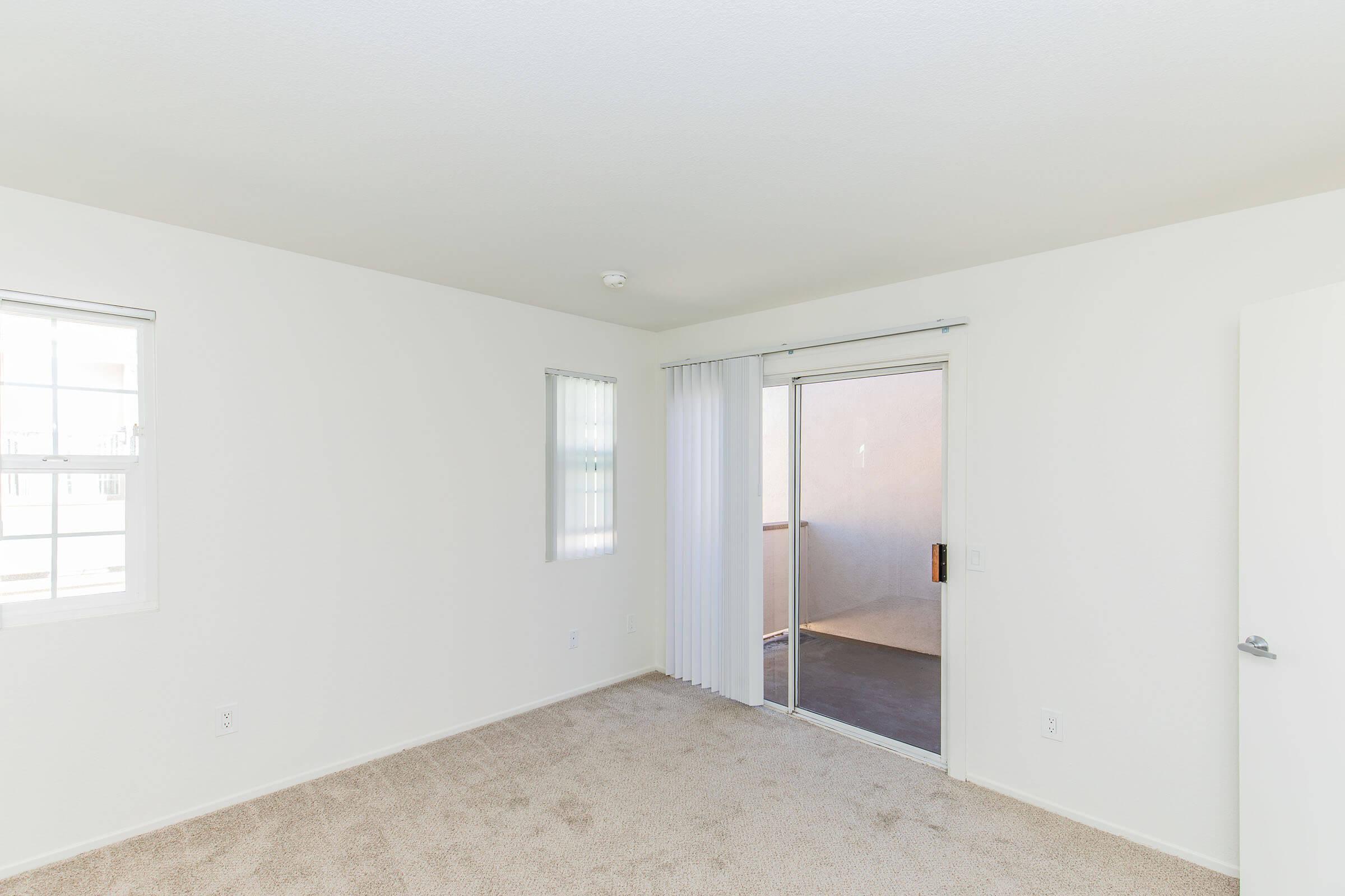 A bright, empty room with light-colored walls and beige carpet. It features two windows and a sliding glass door leading to a balcony. The overall atmosphere is spacious and airy, suitable for various interior designs.
