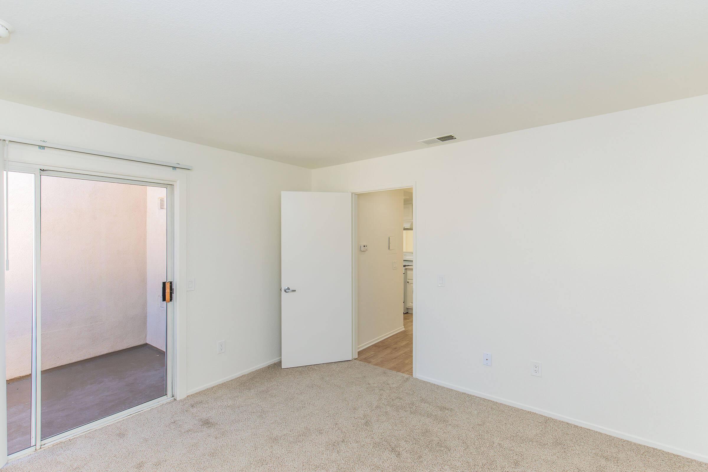 Empty room with light beige carpet and white walls. A sliding glass door leads to a small outdoor area, while a standard interior door is visible on the right, opening to another space. The area is well-lit and spacious, creating an airy feel.