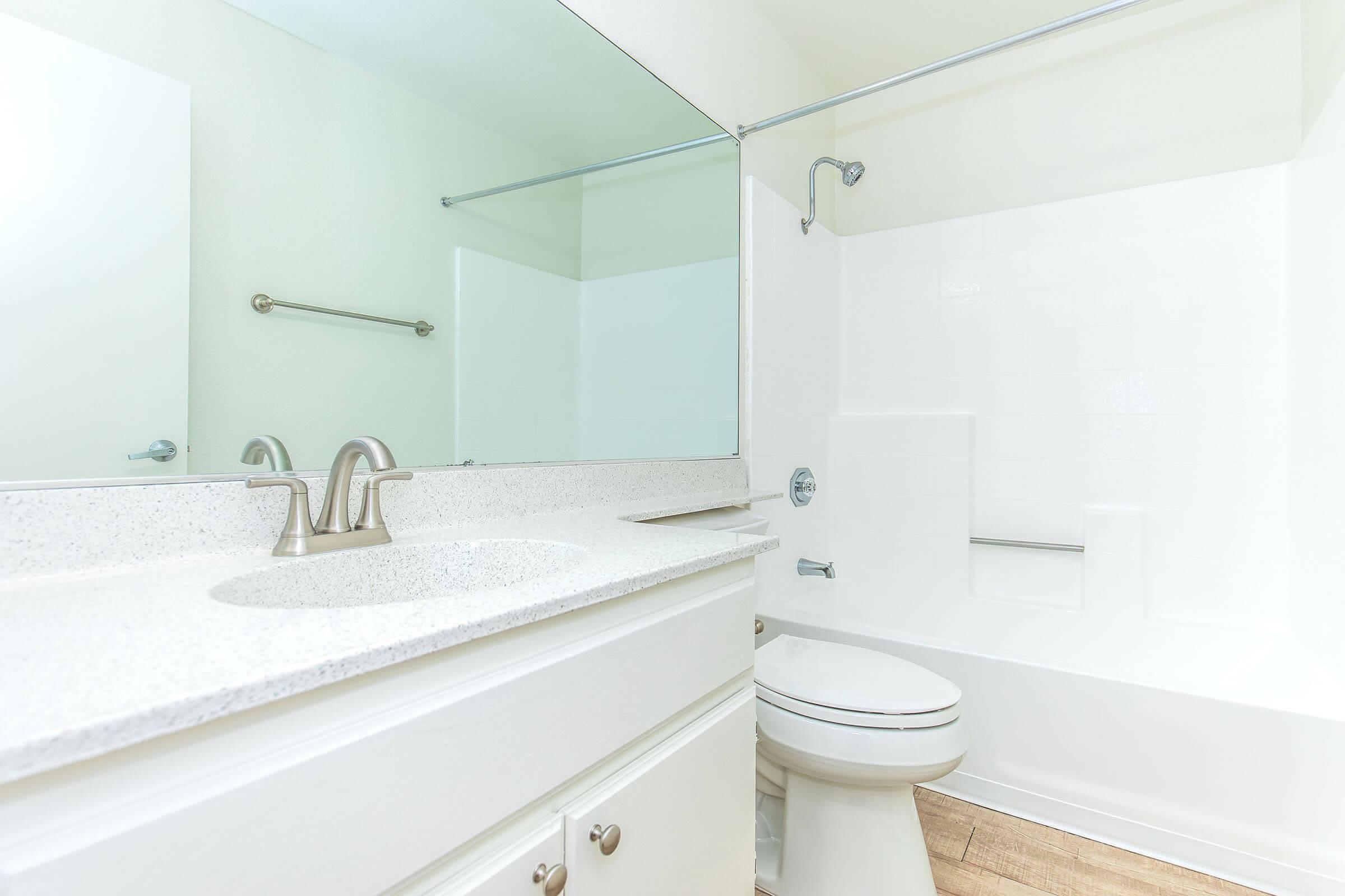 A clean, modern bathroom featuring a white countertop with two faucets, a wall-mounted mirror, a bathtub with a shower, and a white toilet. The walls are light-colored, and there is a towel bar beside the mirror, creating a bright and spacious feel.