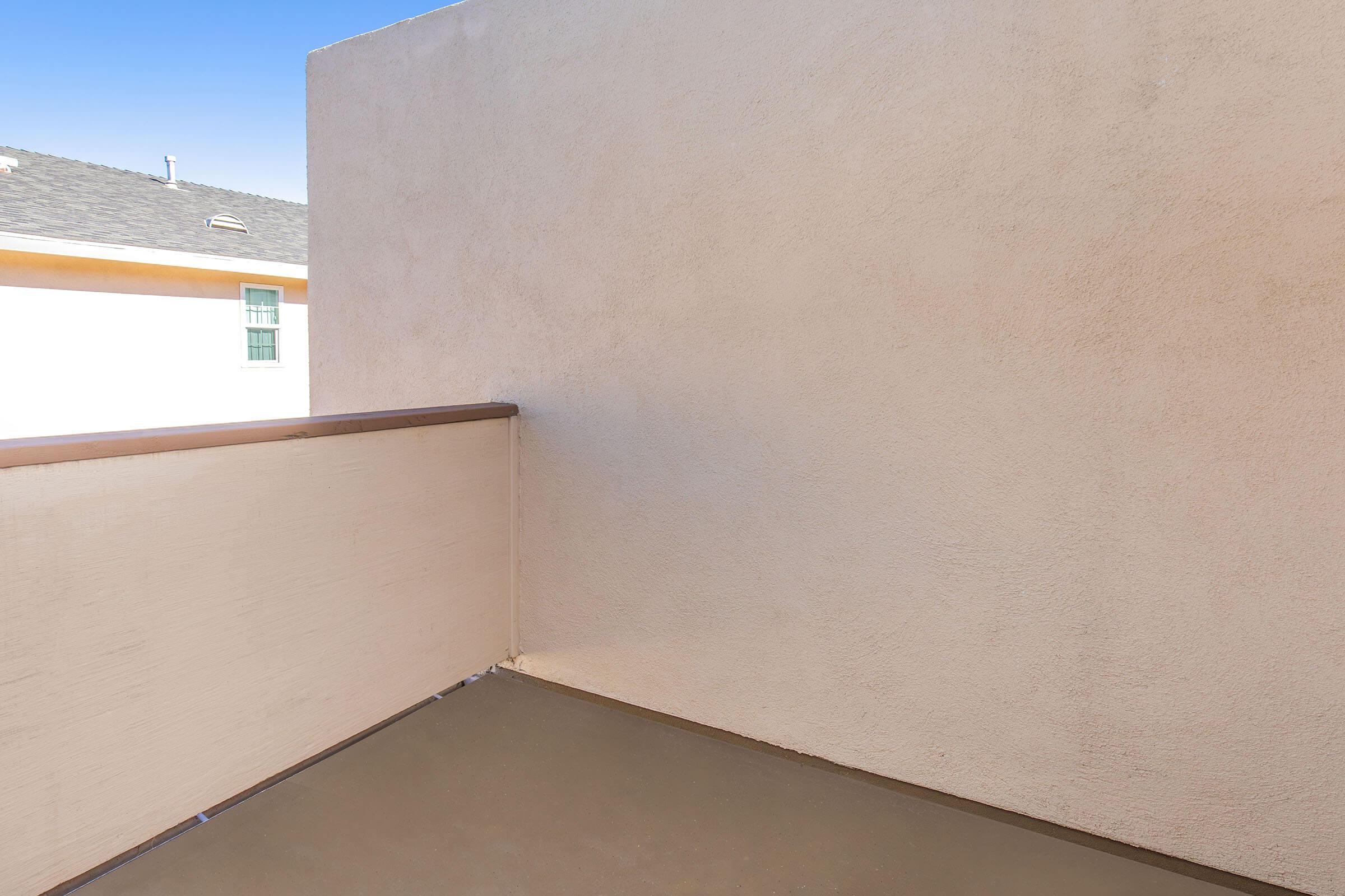 A small balcony view with a textured beige wall on one side and a railing. The floor is smooth and gray, and there's a glimpse of a neighboring building in the background. The sky is clear and bright above, indicating a sunny day.