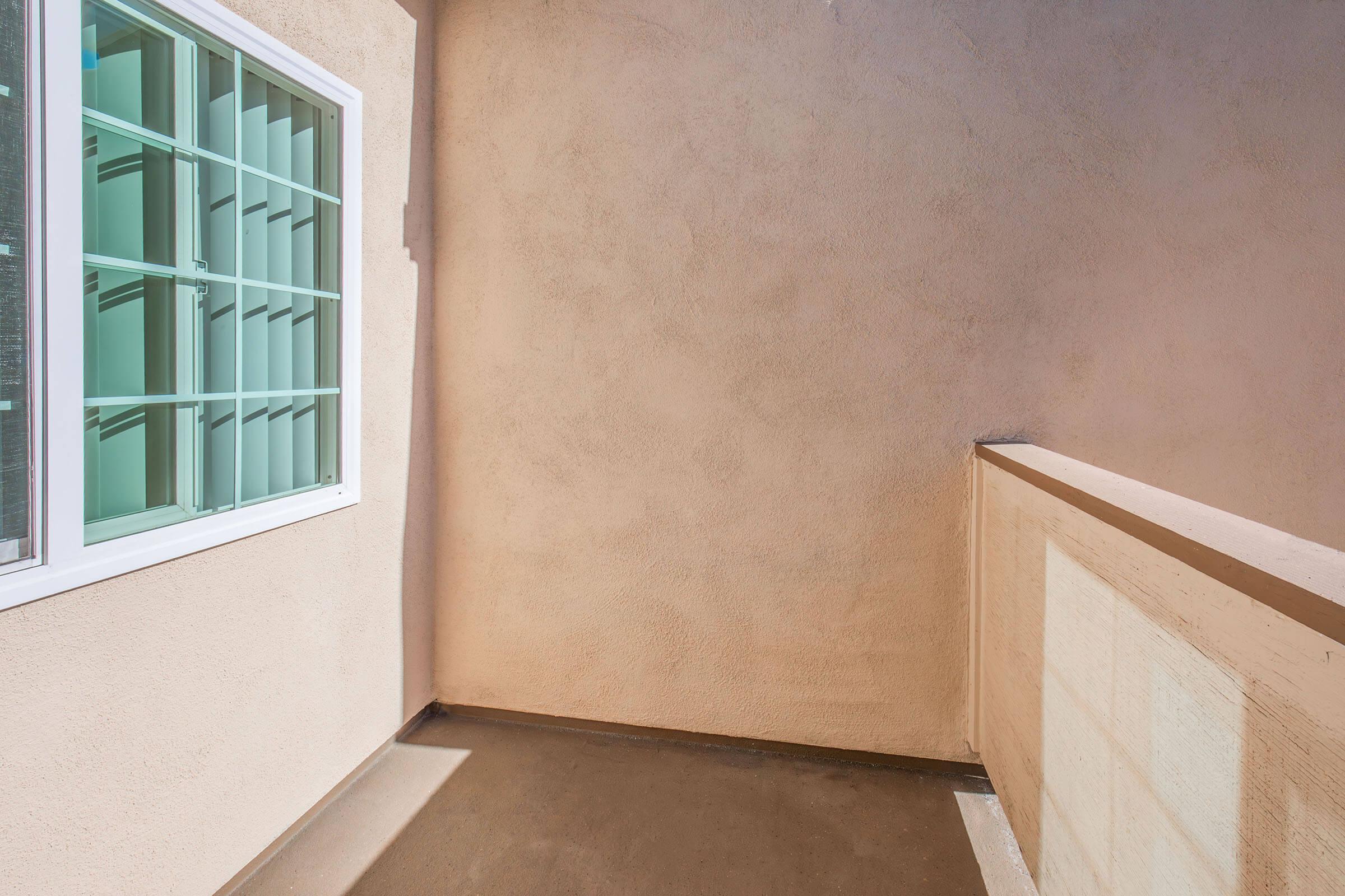 A sunlit corner of a balcony featuring a textured beige wall and a window with a light green frame, casting soft shadows on the floor. The space is minimalistic, creating a serene atmosphere.