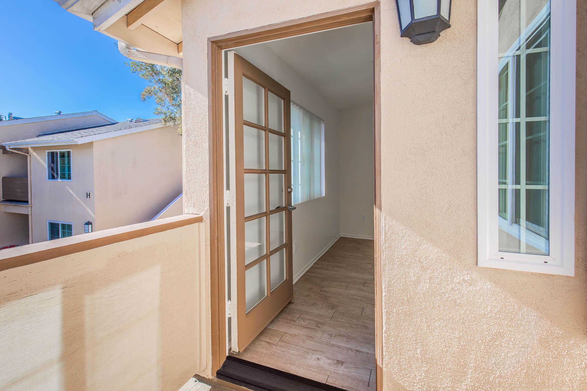 An open door leading to a light-filled room with large windows. The room has a light-colored wooden floor, and the walls are painted in a soft hue. A balcony view shows neighboring buildings and a clear blue sky. A lamp fixture is visible beside the door.