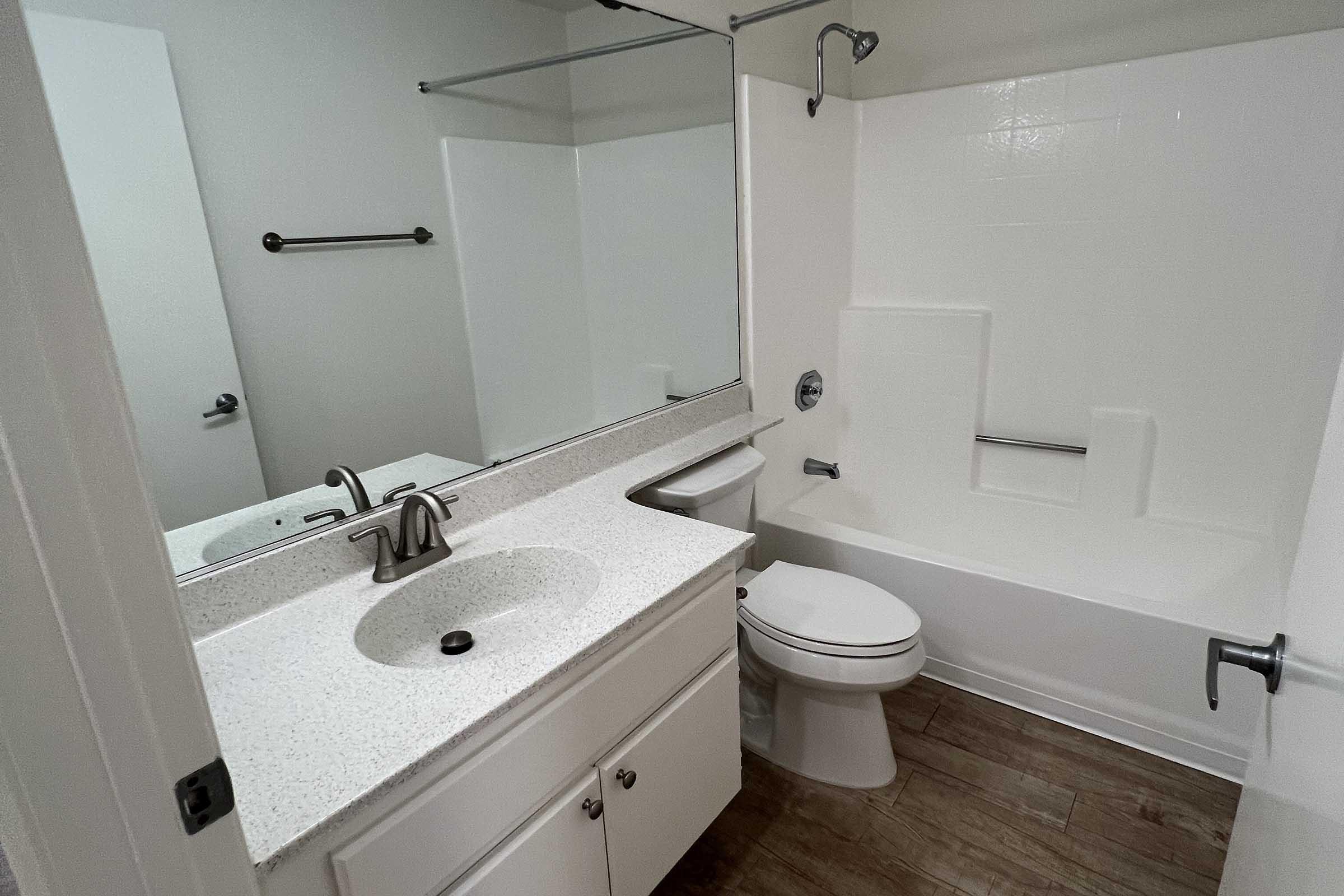 A clean and modern bathroom featuring a white bathtub with a shower, a sleek vanity with a sink, a large mirror, and wooden flooring. The space includes a toilet and is well-lit, creating a bright and airy atmosphere.