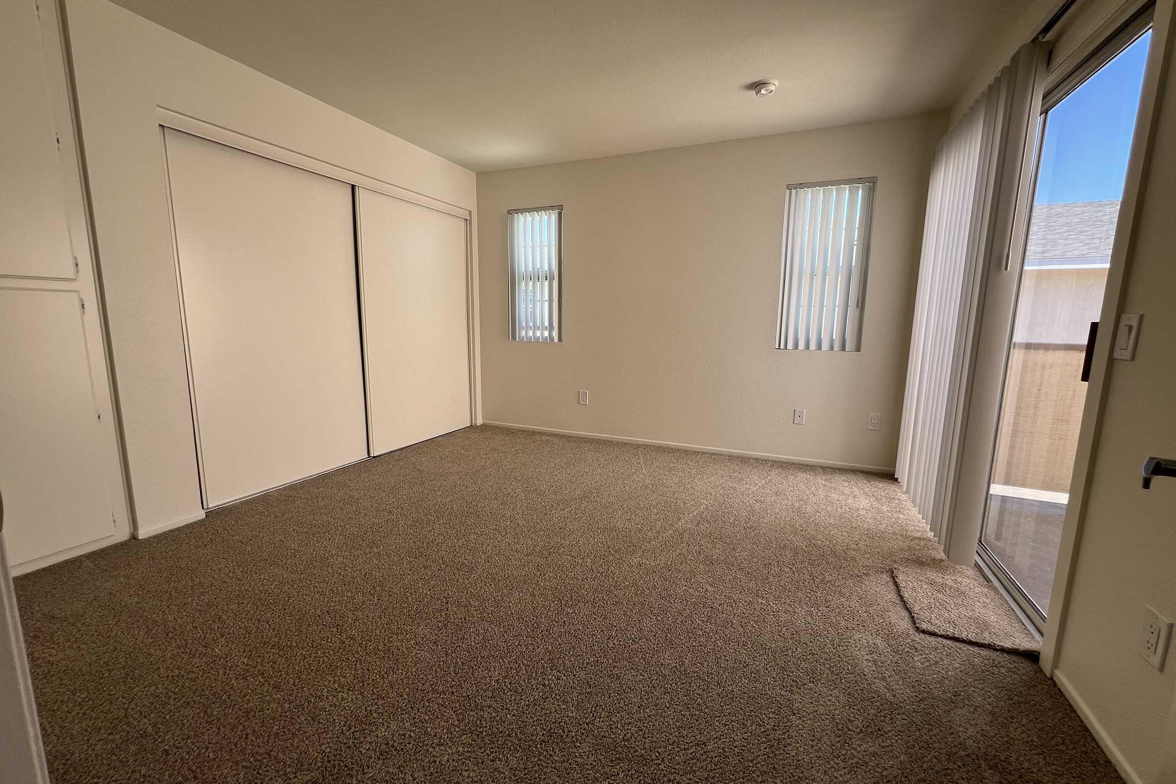 A spacious, empty room with beige carpet and white walls. It features sliding closet doors, two narrow windows with blinds, and a glass sliding door leading to an outdoor area. The overall atmosphere is bright and airy, suitable for various uses.