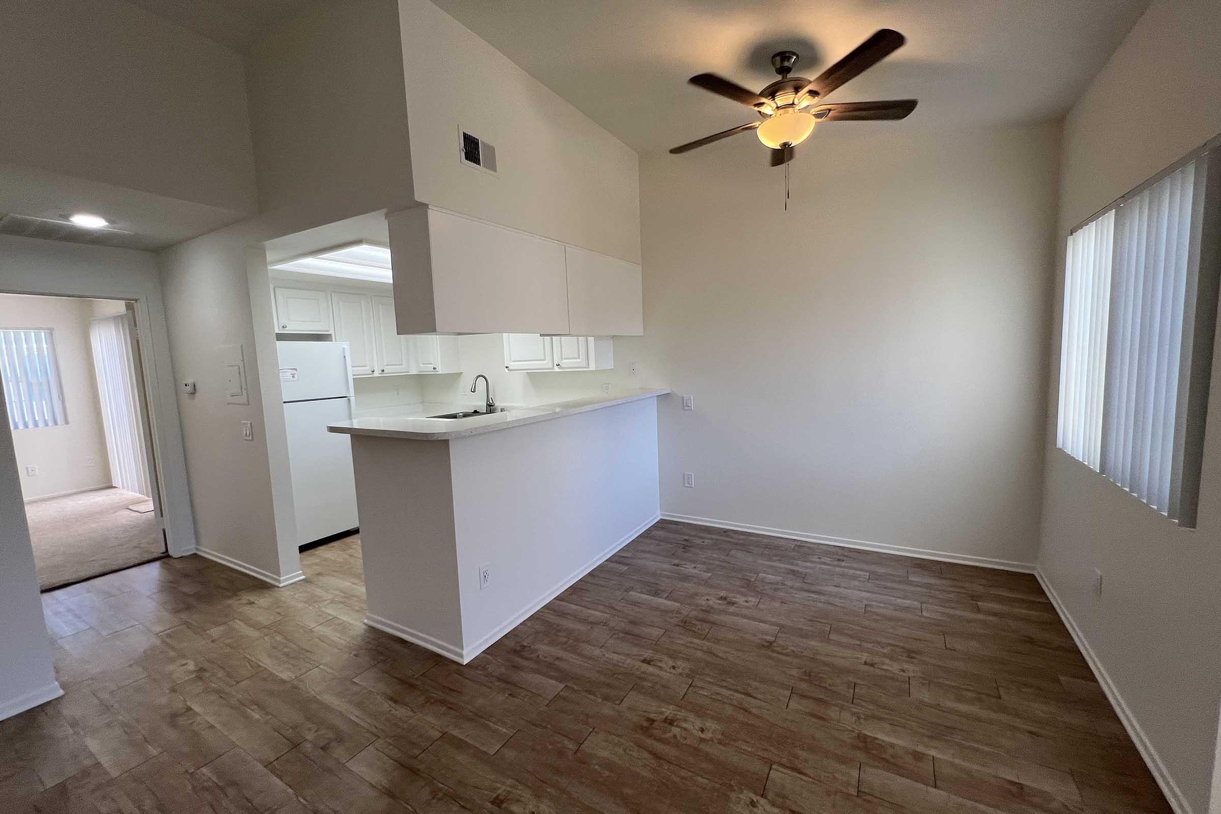 Bright and airy interior of an apartment featuring a kitchen area with a bar counter, a ceiling fan, and a living space with wood-like flooring. The walls are painted in a light color, and windows allow natural light to fill the room. Adjacent is a separate room with carpet flooring.