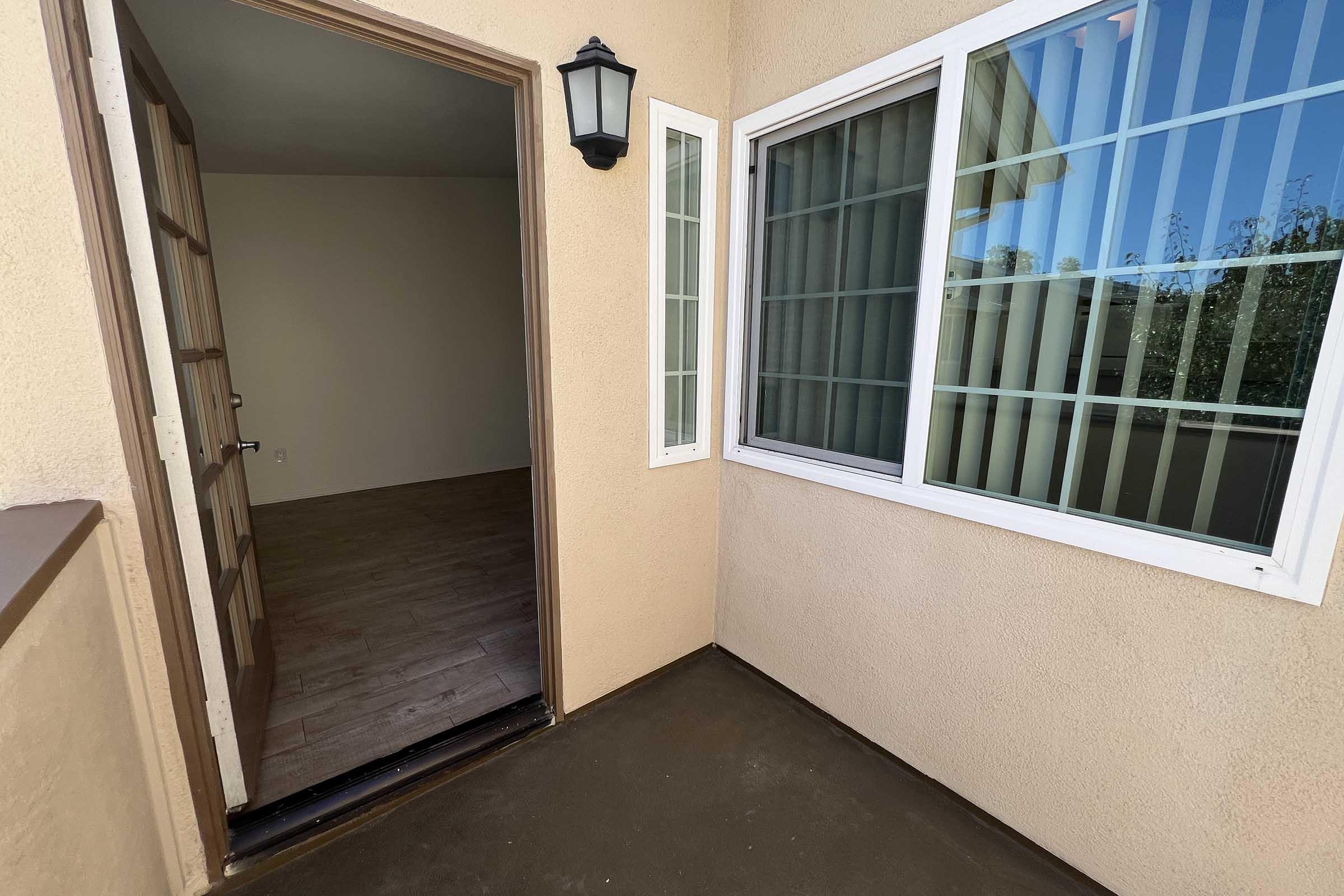 A view of an open doorway leading into an empty room with light-colored walls and a wooden floor. A lantern light is mounted next to the door, and there is a large window with vertical blinds visible to the right. The area outside the door is clean and well-lit, suggesting a welcoming entrance.