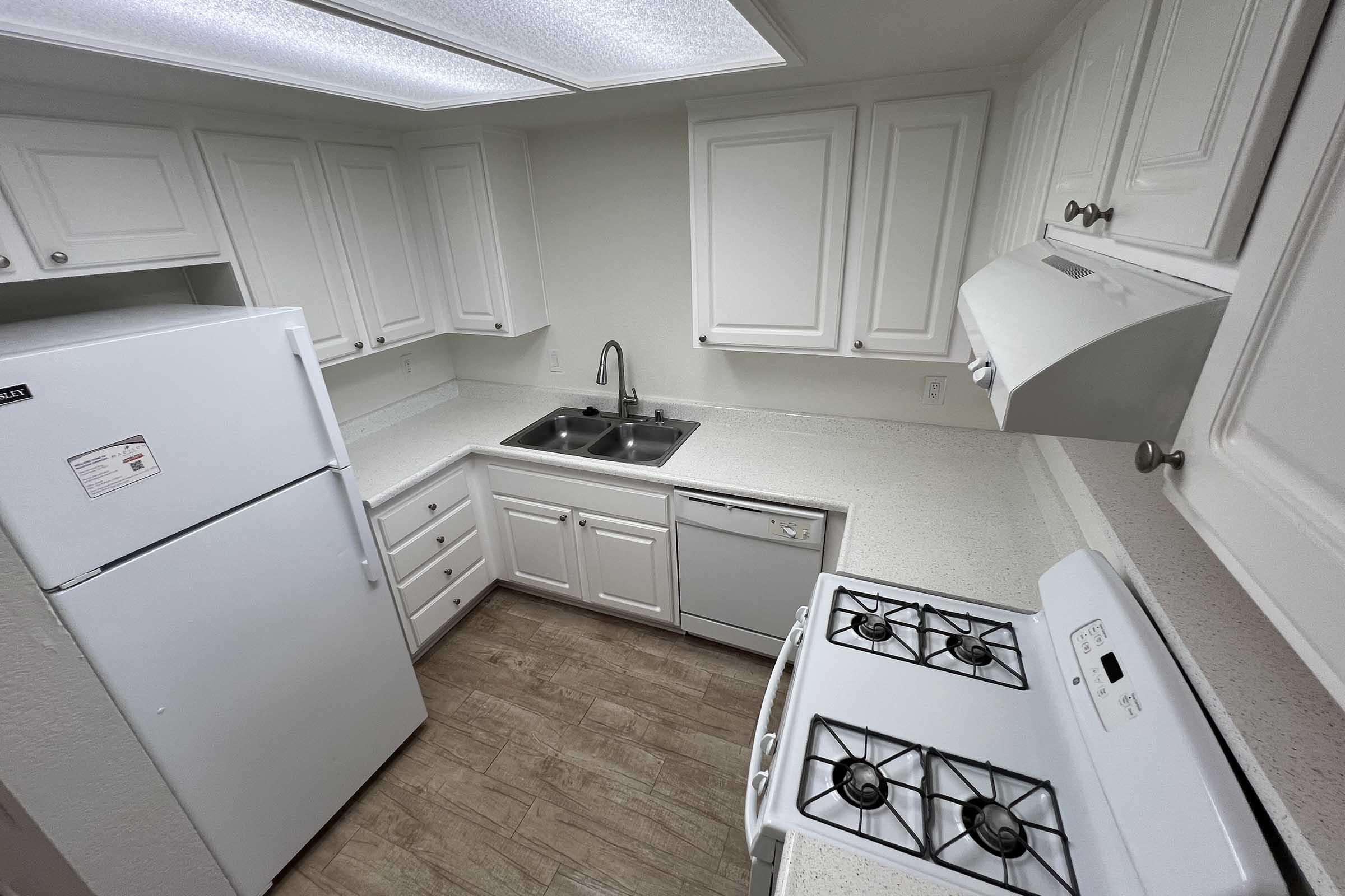 A clean and modern kitchen featuring white cabinets, a refrigerator, a stove with an oven, and a double sink. The countertops are light-colored, and there is a window above the sink. The floor is a wood-like laminate, giving the space a bright and inviting atmosphere.