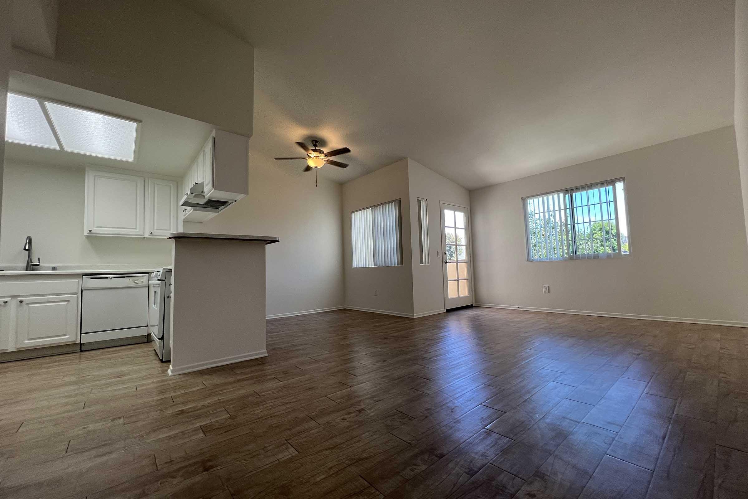 Bright and spacious living area featuring a ceiling fan, large windows allowing natural light, and a view of an outdoor balcony. The adjoining kitchen includes white cabinetry and modern appliances. The flooring is light-colored wood-like laminate, giving a warm and inviting atmosphere.