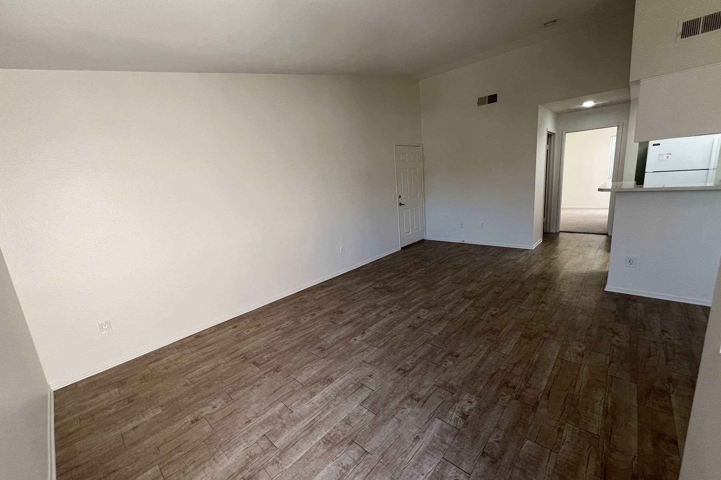 A spacious, unfurnished room with light-colored walls and a wooden floor. There's a door on the left and an opening leading to a hallway in the background. Natural light enters from a doorway in the distance, creating a bright atmosphere. The layout is simple, ideal for customizing with furniture.
