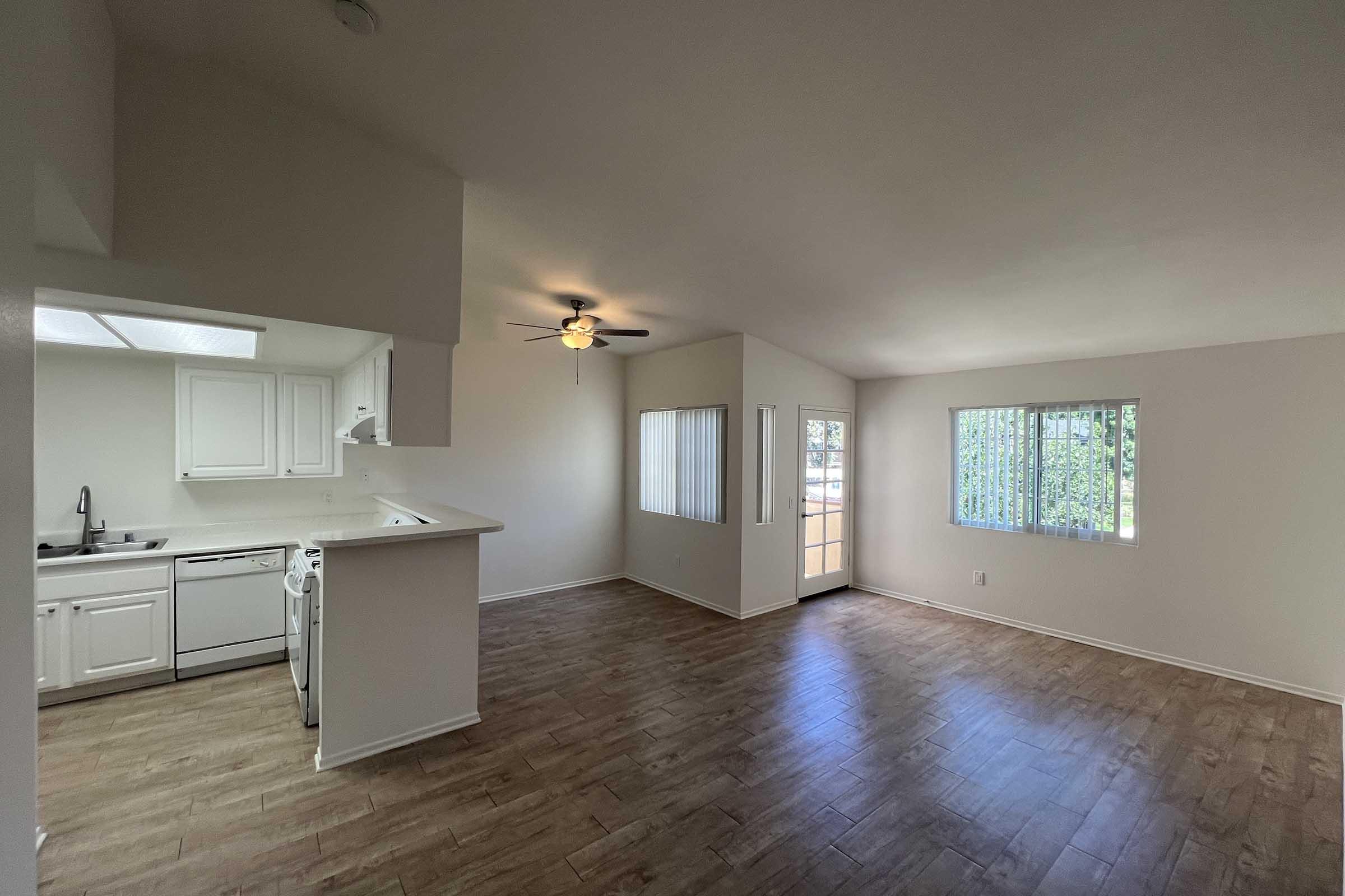 Spacious interior of an apartment featuring a living area with a ceiling fan, large windows allowing natural light, and an open kitchen with white cabinetry and appliances. The flooring is a light-colored wood, and there is a door leading to an outdoor area.