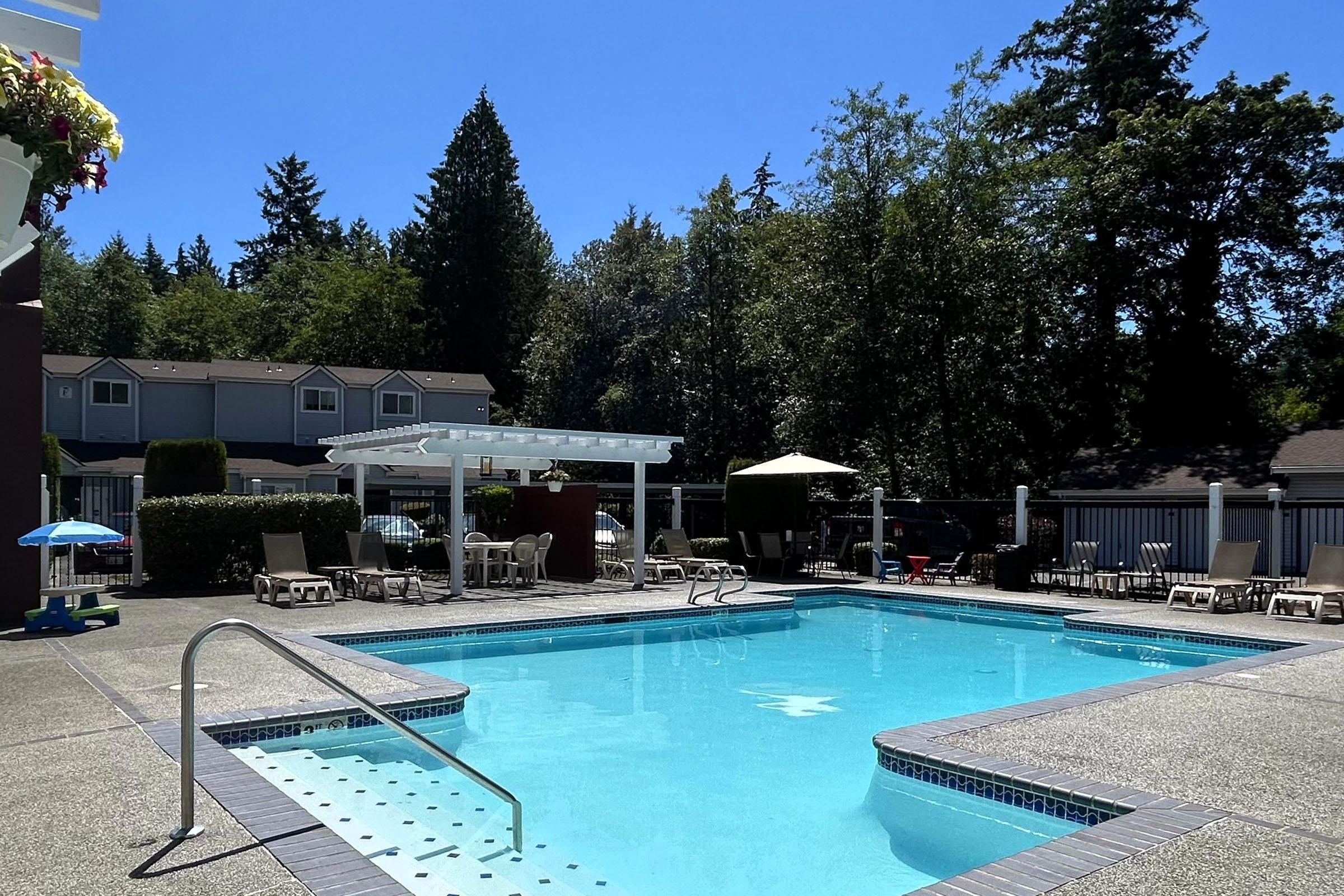 A tranquil outdoor swimming pool surrounded by lounge chairs and umbrellas. Lush green trees provide a scenic backdrop, and a pergola offers shaded seating. Clear blue sky and sunlight create a relaxing atmosphere perfect for leisure and enjoyment.