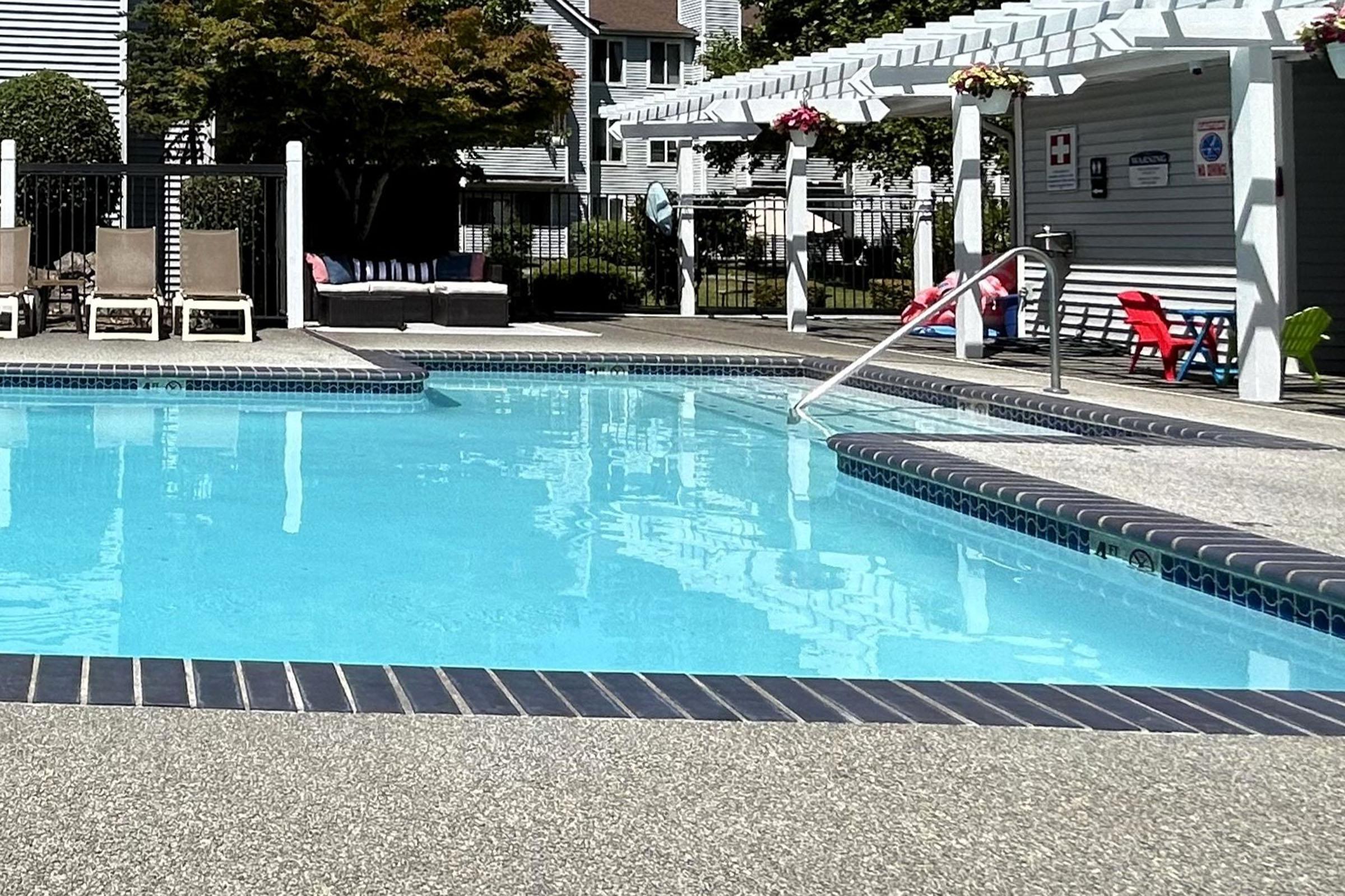 A clear swimming pool with light blue water surrounded by a textured concrete deck. Lounge chairs are arranged nearby, and a shaded area features seating. In the background, there are houses and greenery, creating a relaxing outdoor atmosphere.