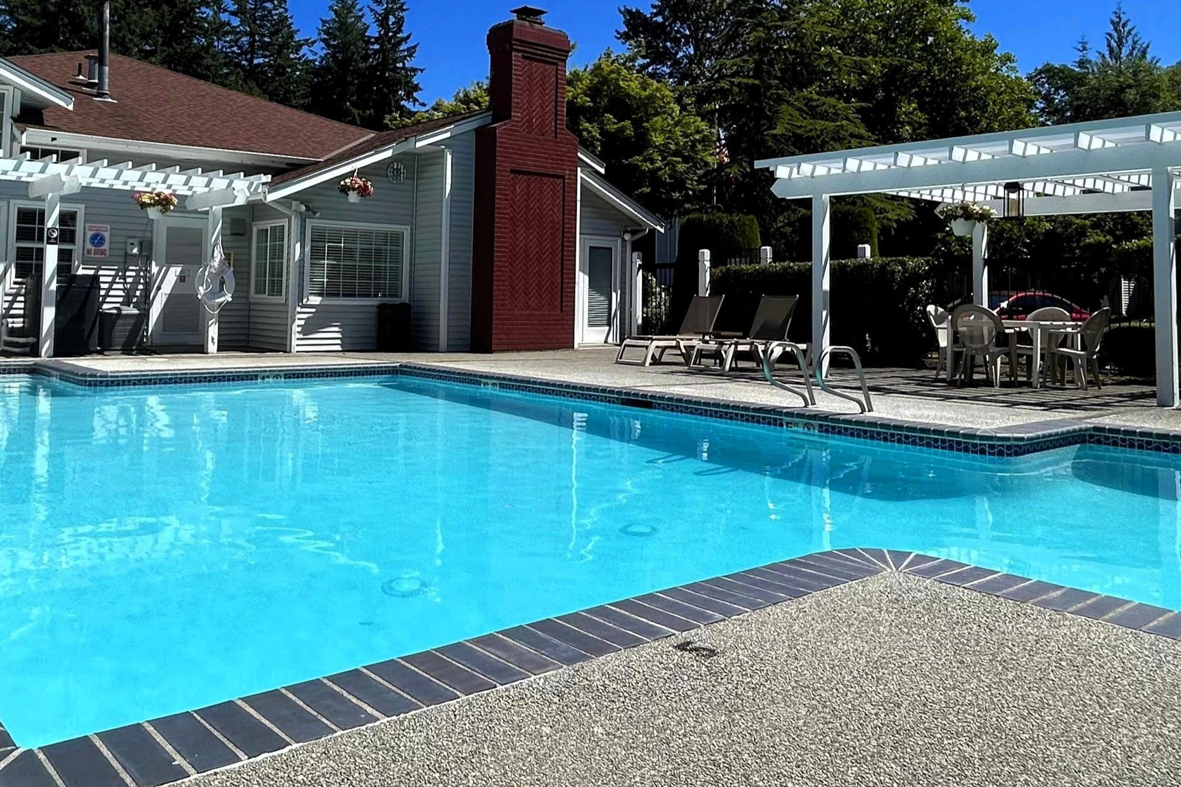 A bright outdoor swimming pool with clear blue water, surrounded by lounge chairs and a dining table under a pergola. In the background, a building with a chimney and lush greenery offers a relaxing atmosphere. Sunlight casts a warm glow over the scene, creating an inviting setting for leisure.