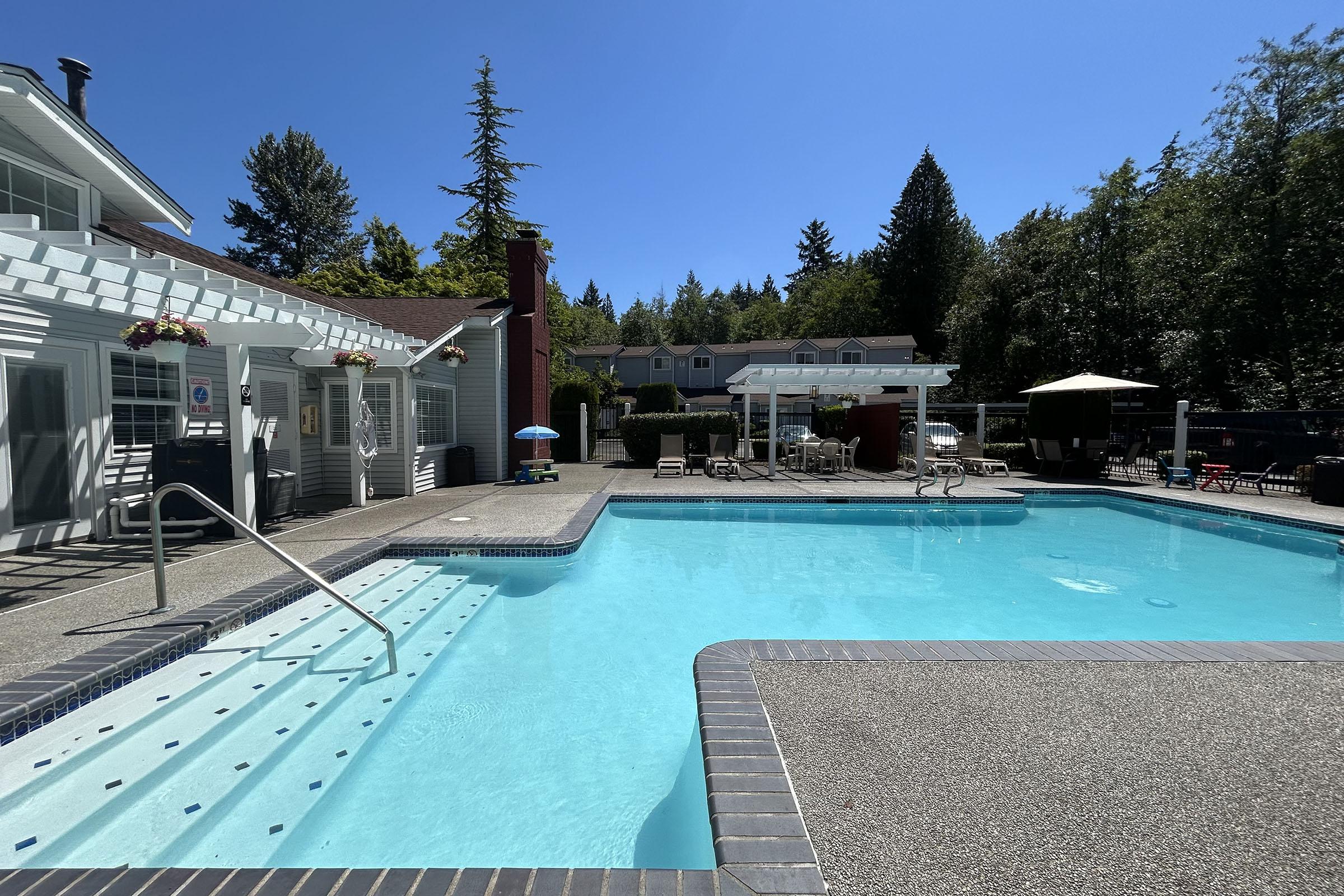 A bright, clear swimming pool surrounded by a patio area with lounge chairs and umbrellas. In the background, there are trees and additional buildings. The sky is blue and sunny, creating a relaxing outdoor atmosphere.