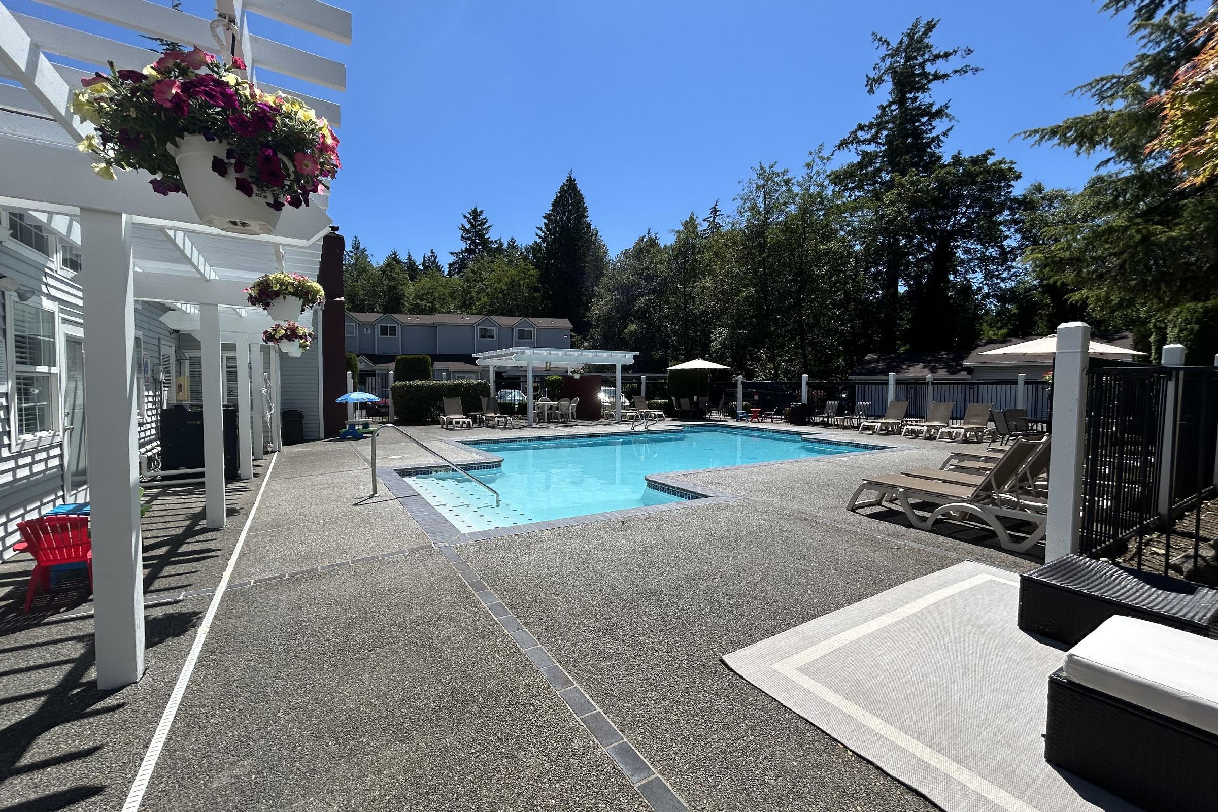 A sunny outdoor swimming pool area surrounded by lounge chairs and umbrellas. The pool is inviting, with clear blue water, situated in a landscaped setting with trees in the background and flower pots hanging on the railing. An area of relaxation is visible nearby.