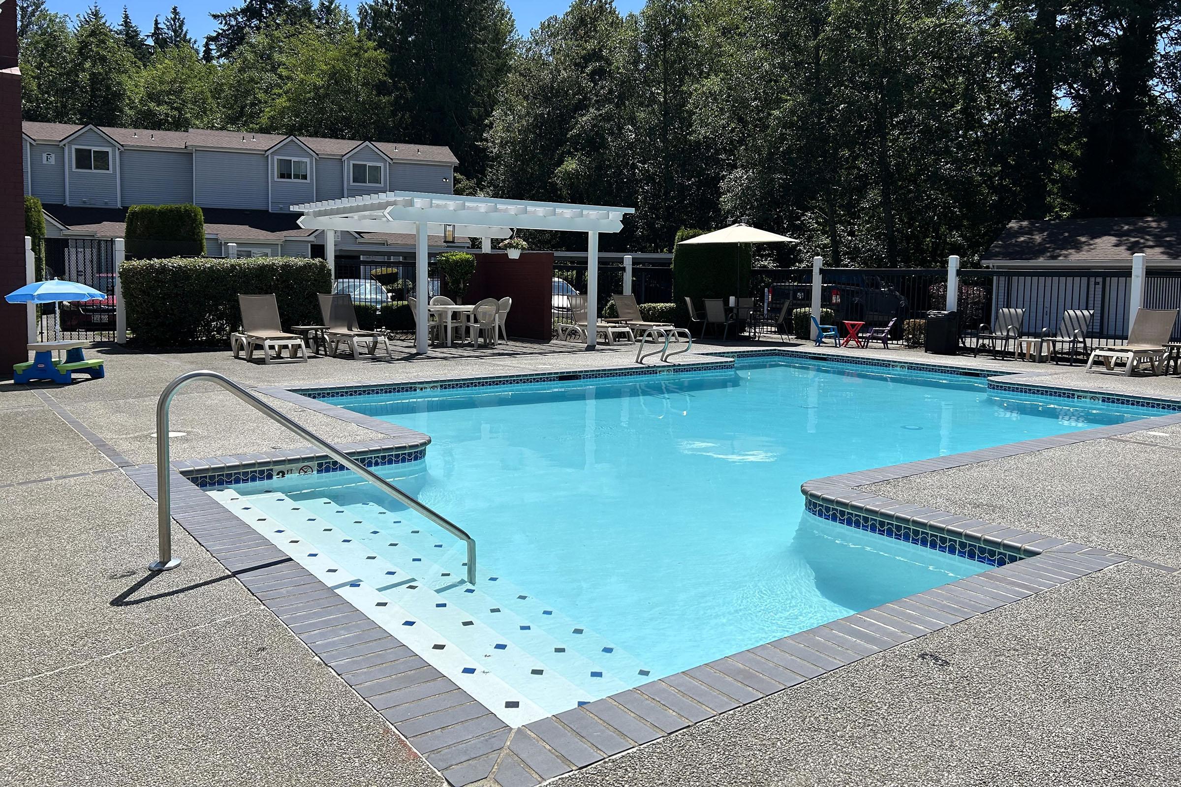 A clear swimming pool surrounded by lounge chairs and patio umbrellas, set in a shaded area with trees in the background. There is a pergola providing shade and a children's play area nearby. The pool area features a textured concrete deck with decorative tiles along the edge.