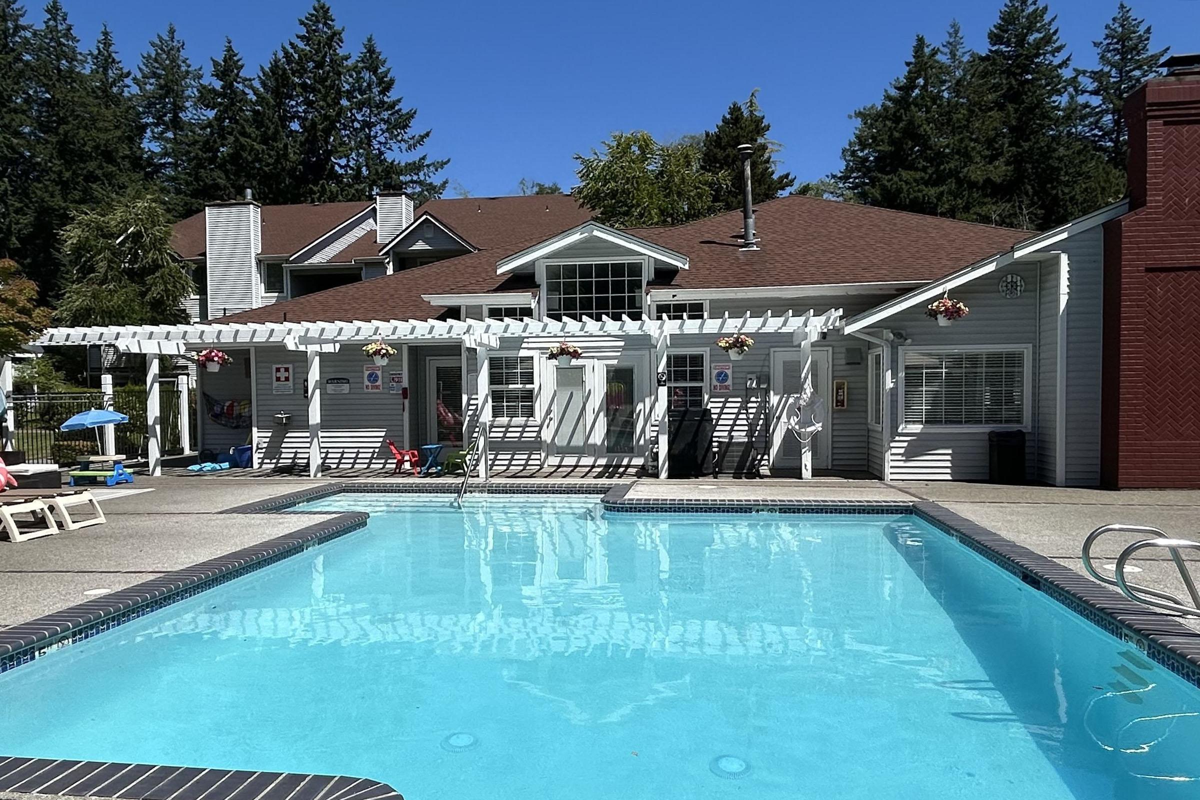 A sunny outdoor swimming pool area with a clear blue pool in the foreground. Behind it is a single-story building with a sloped roof, large windows, and a covered patio. Surrounding the pool are lounge chairs, umbrellas, and colorful decorations, with tall trees in the background.