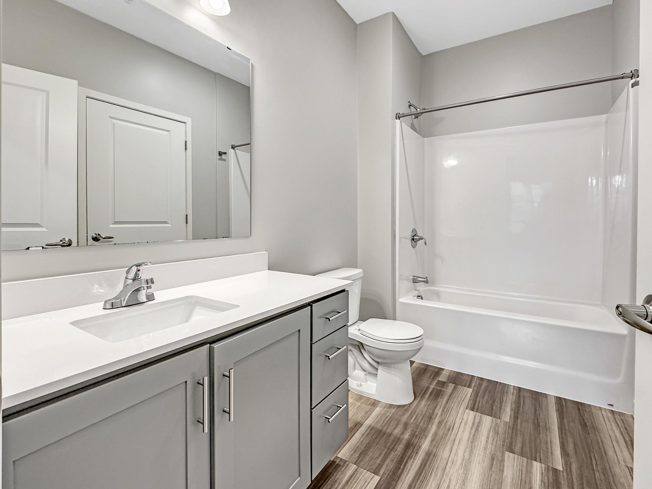 Modern bathroom featuring a double sink vanity with gray cabinetry, a large mirror, and a white countertop. The space includes a toilet and a bathtub with a shower curtain. The floor has wood-like tiles, and the walls are painted a neutral gray. Bright lighting enhances the clean, contemporary look.