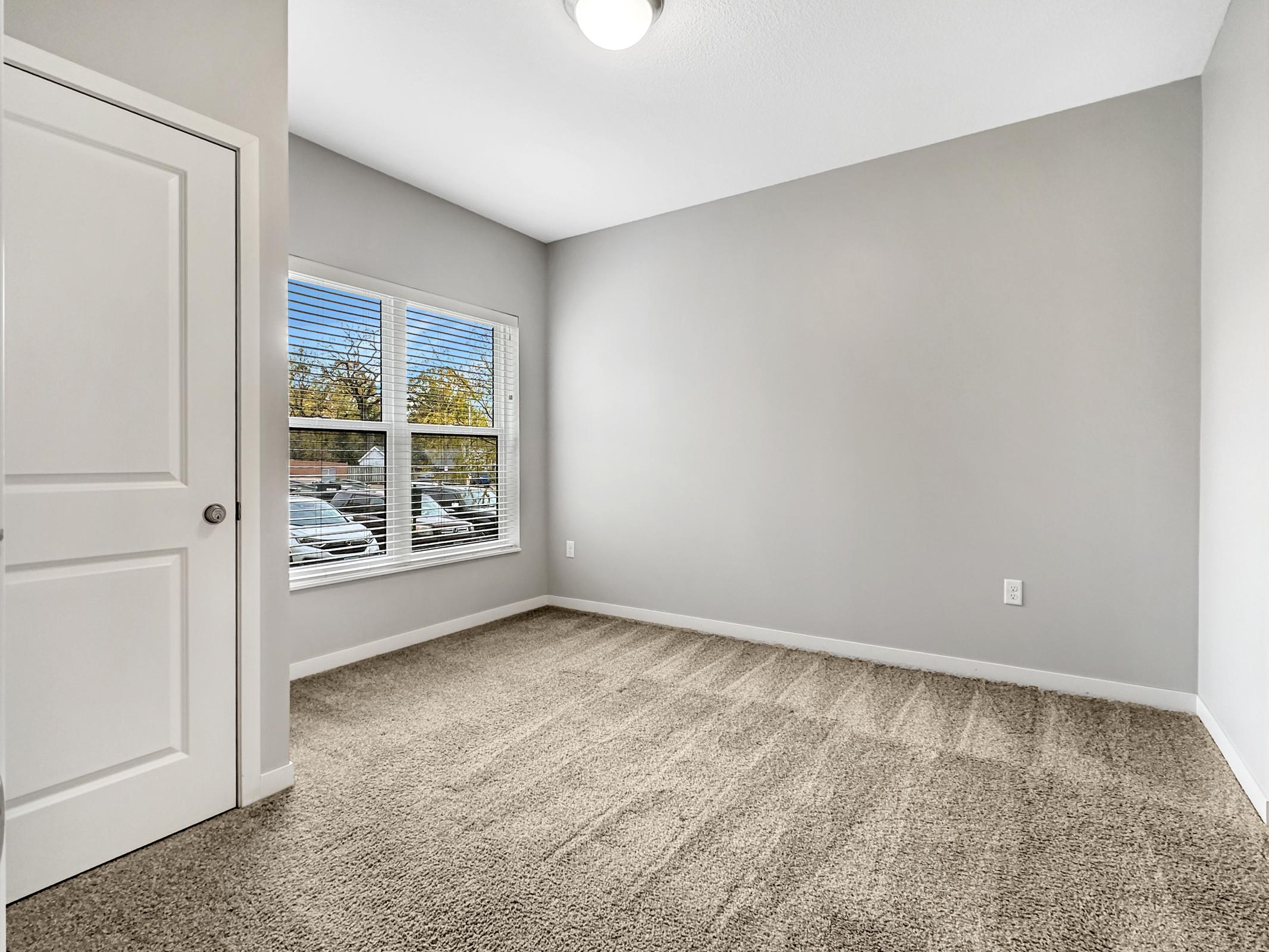 A bright, empty room with gray walls and a carpeted floor. A large window lets in natural light, revealing a view of parked cars outside. The room features a white door and simple lighting, creating a clean and minimalistic atmosphere.