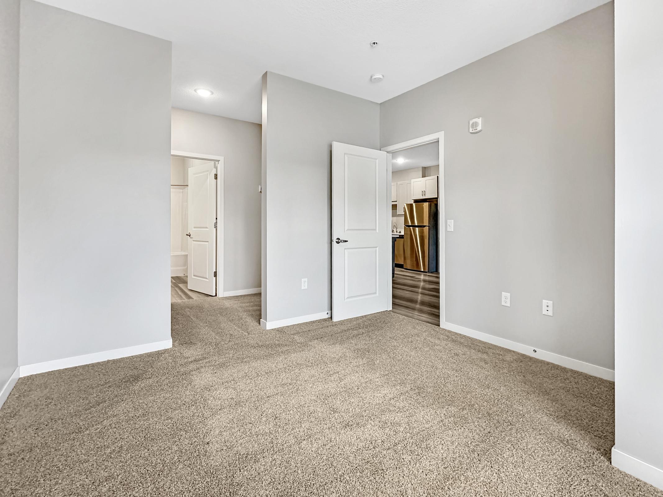Interior view of a spacious room with light gray walls and plush carpeting. A partially open door leads to another room. In the background, a glimpse of a kitchen area is visible. The lighting is bright and the atmosphere is modern and inviting.