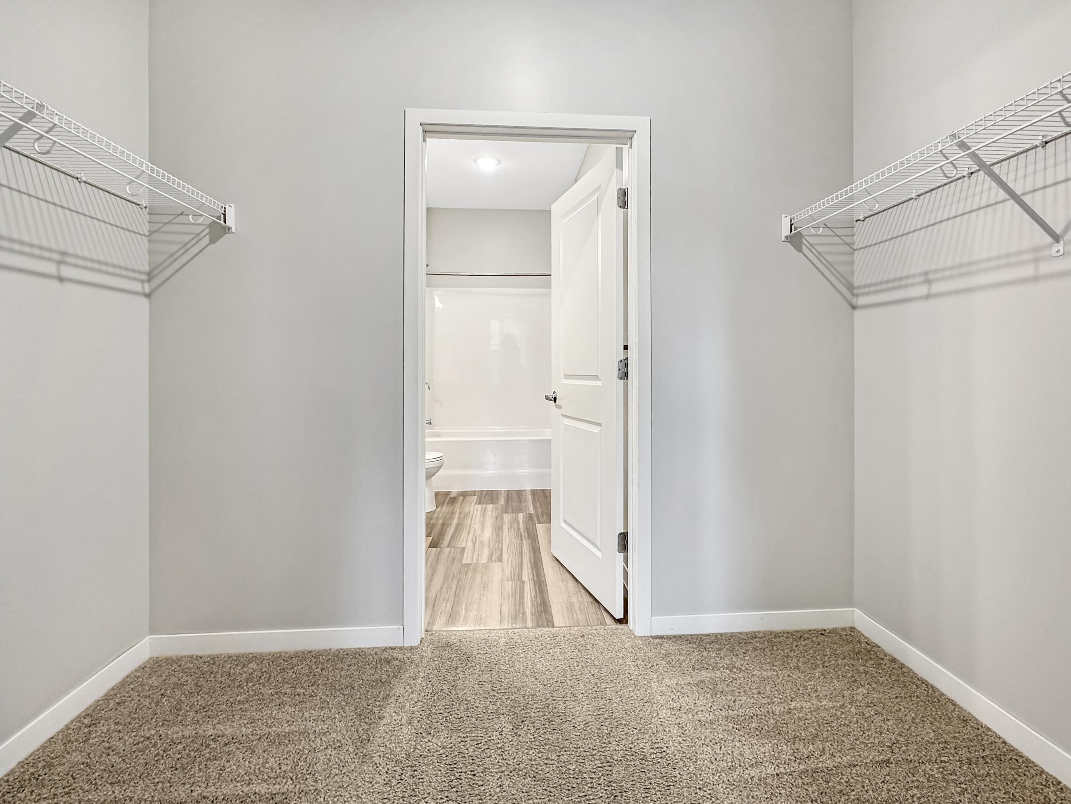 A spacious empty closet with light gray walls, featuring two metal wire shelves on opposite sides. A doorway leads to a bathroom with white walls and a light-colored floor. The closet floor is covered in beige carpet, creating a clean and open atmosphere.