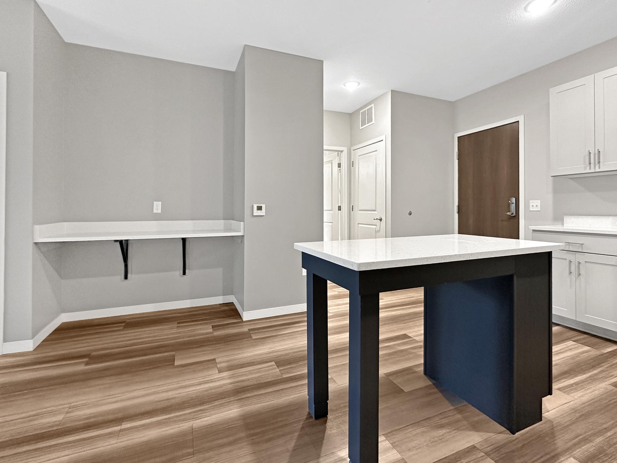 A modern kitchen interior featuring light-colored walls, wooden flooring, and a central island with a dark base. There is a small countertop area along one wall, and a door leading to another room in the background. The overall design is sleek and contemporary.