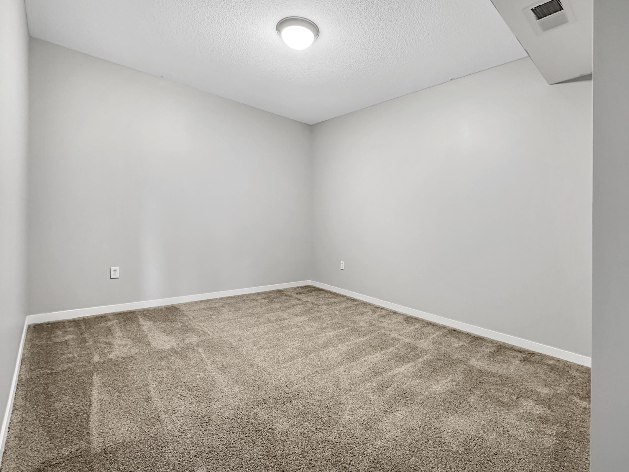 A vacant room with light gray walls and a ceiling light. The floor is covered in beige carpet, and there are no furnishings. The space appears clean and well-lit, with a simple design and minimal decor.