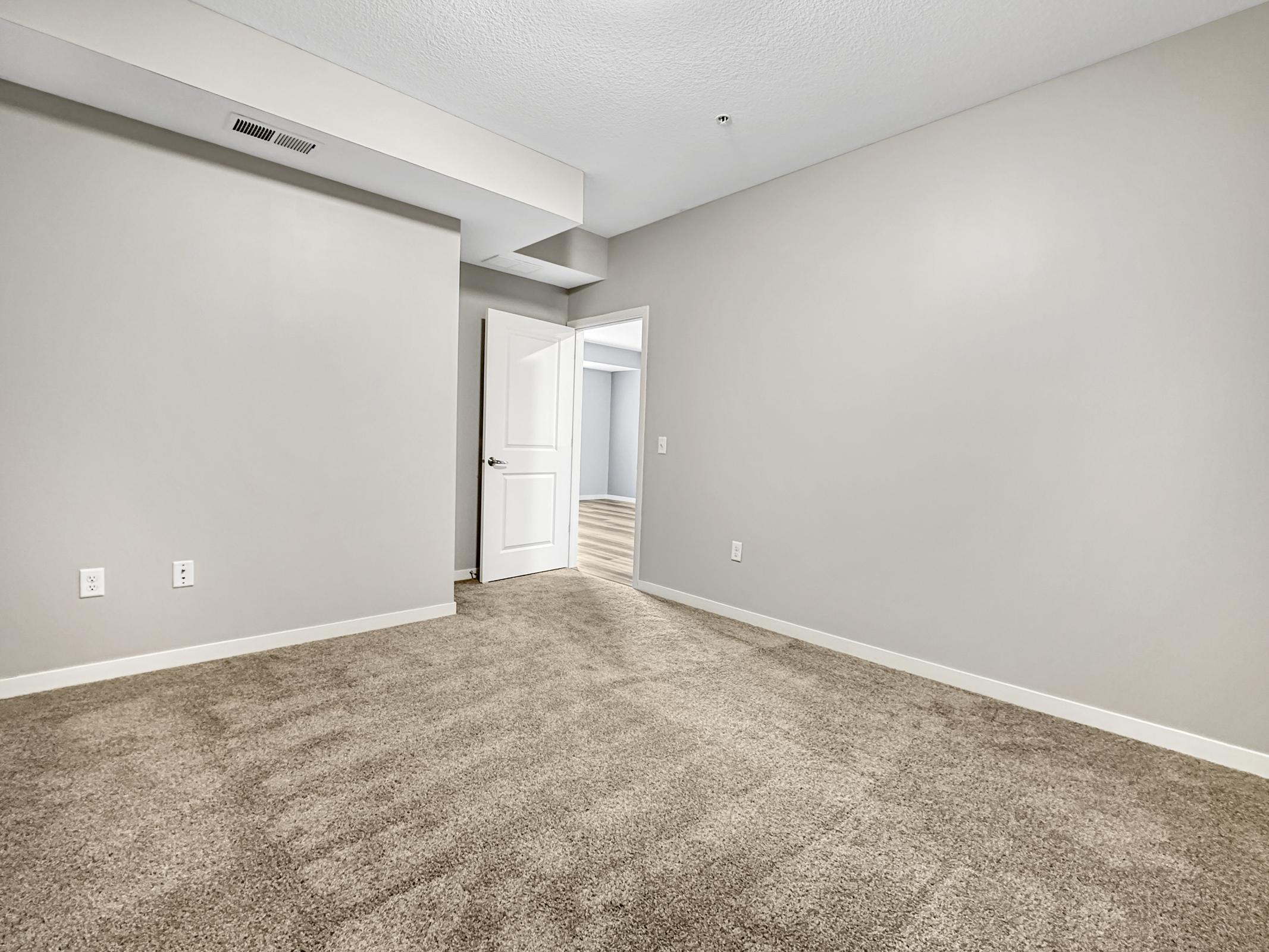 Empty room with gray walls and a light beige carpet. A doorway leads to another room, and there's a white door partially opened in the background. The space is well-lit and features a neutral decor, creating a blank canvas for interior design.