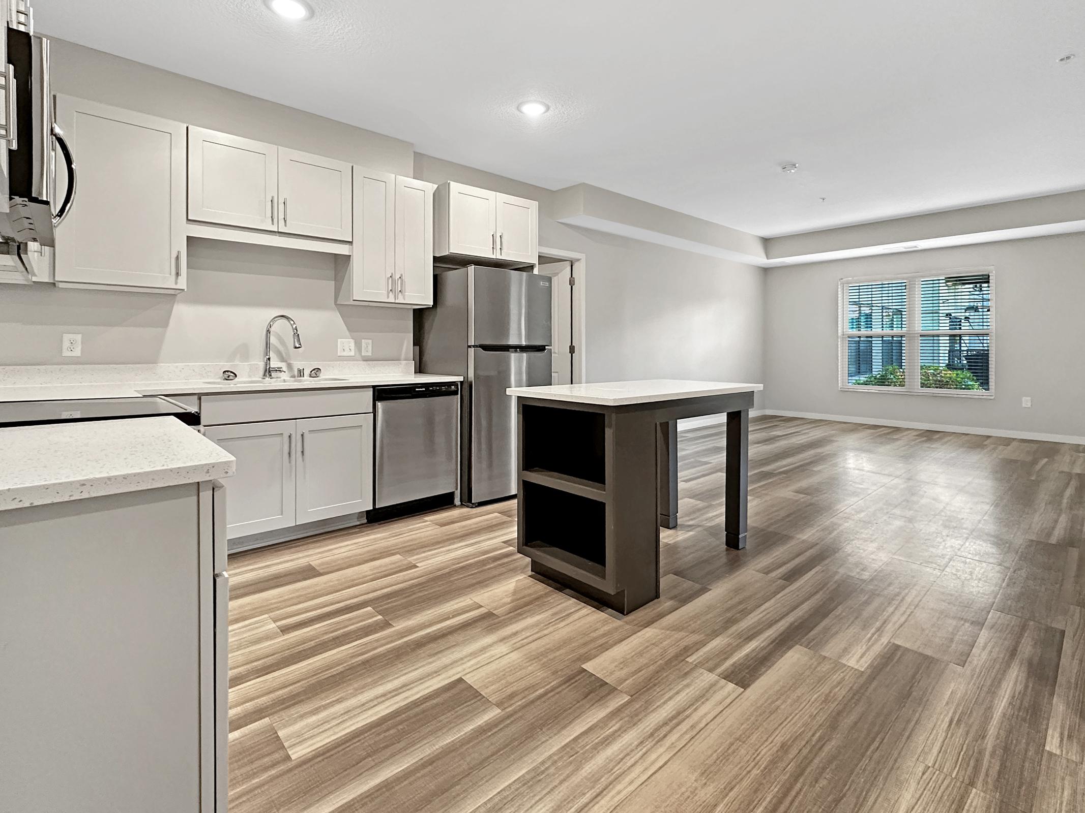 Modern kitchen and living area featuring sleek cabinetry, stainless steel appliances, and an island. The open layout highlights spacious wood-patterned flooring, with natural light streaming through a window in the background. Ideal for cooking and entertaining.