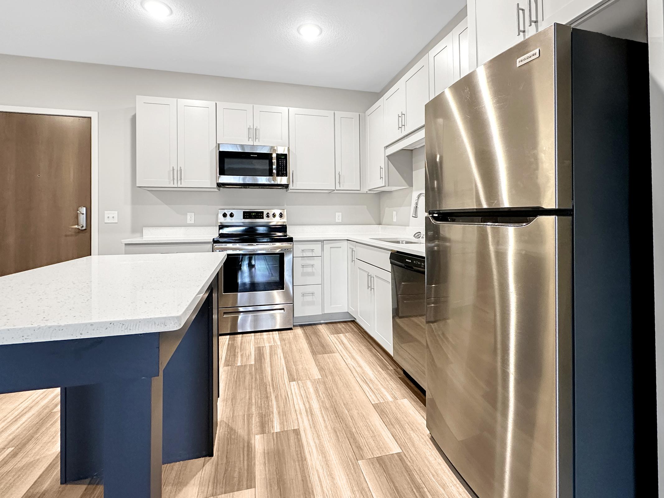 Modern kitchen featuring stainless steel appliances, including a refrigerator, microwave, and oven. The cabinetry is sleek and white, complemented by a light-colored countertop. A central island provides additional counter space, and the flooring is a warm wood-tone laminate. Bright lights illuminate the space.