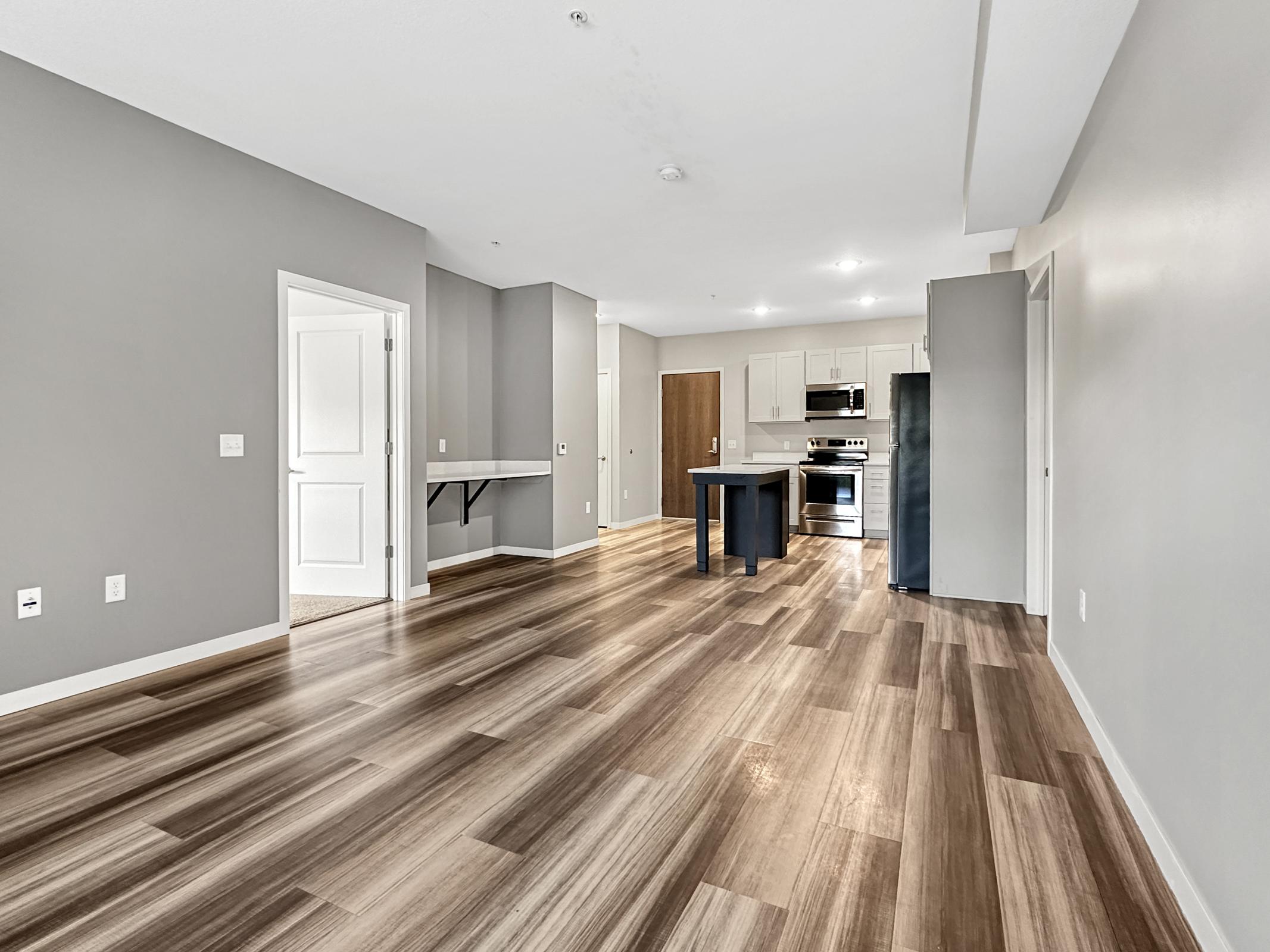 A modern, open-concept living space featuring light gray walls, beautiful wood-like flooring, and white cabinetry. The kitchen includes stainless steel appliances, an island with seating, and a separate area for a dining table. Natural light fills the room from large windows.