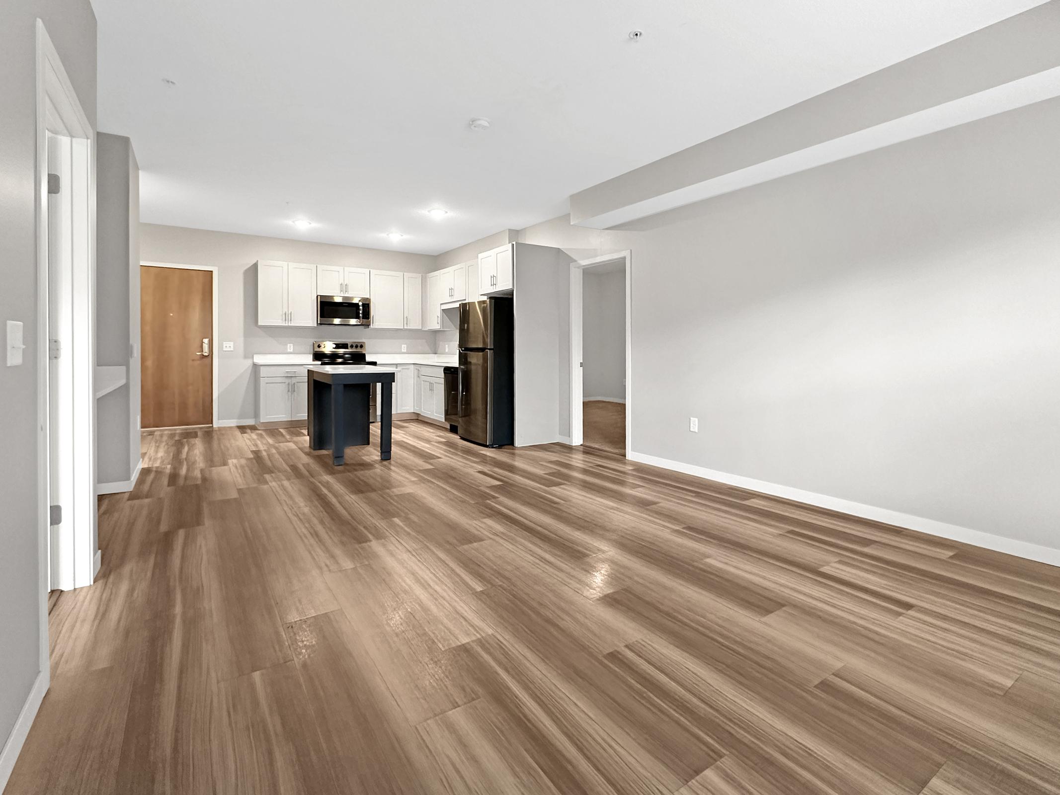 A modern, spacious kitchen and living area with light-colored cabinets, stainless steel appliances, and an island. The floor features a warm, wood-like finish. Bright, neutral walls create an inviting atmosphere. The layout includes an open space with doors leading to other rooms.