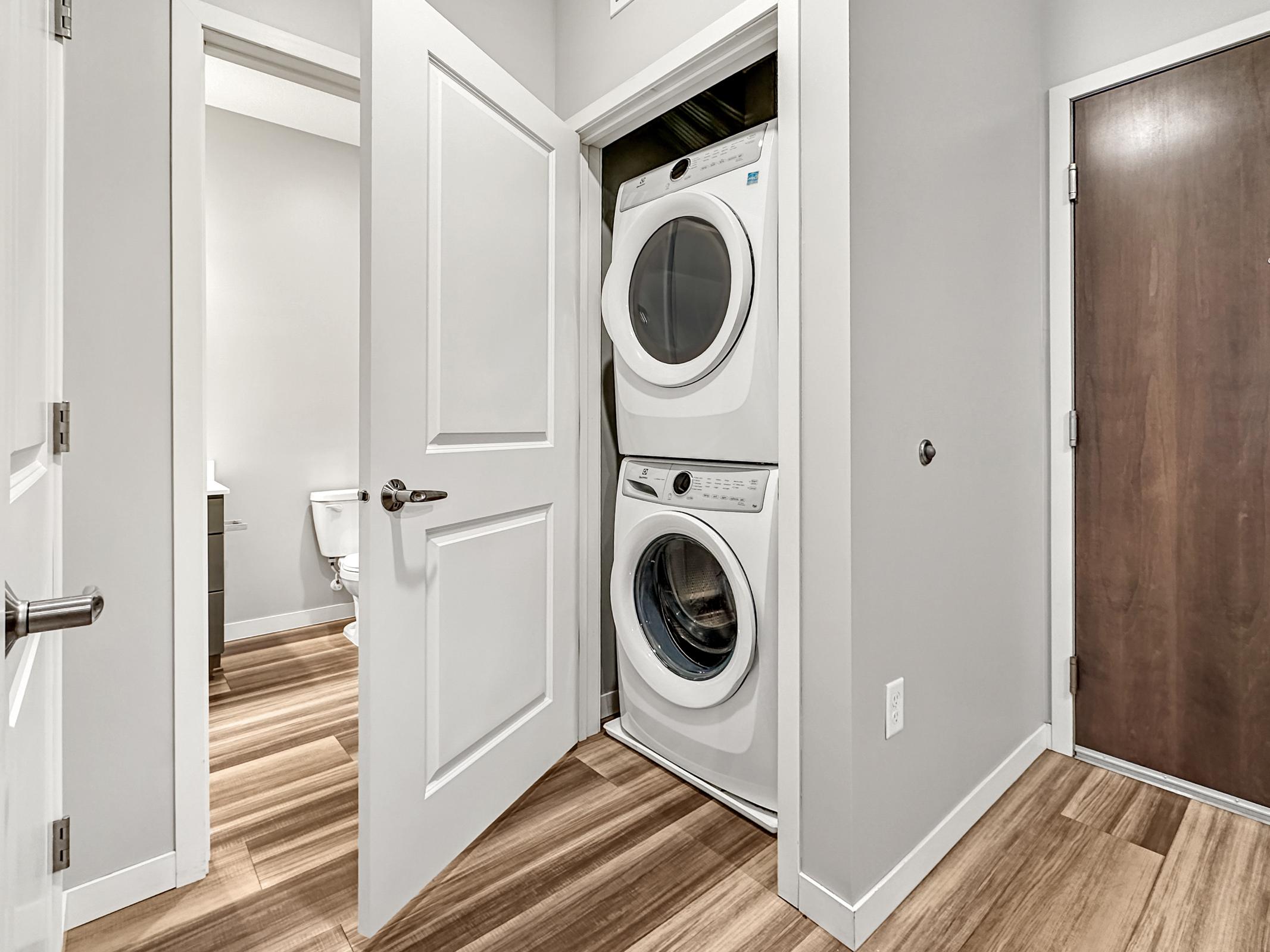 A laundry area featuring a stacked washer and dryer, located in a hallway. The door is open, revealing a light-colored room with gray walls, modern fixtures, and light wooden flooring. A closed door is visible on the right, possibly leading to a bathroom or another room.