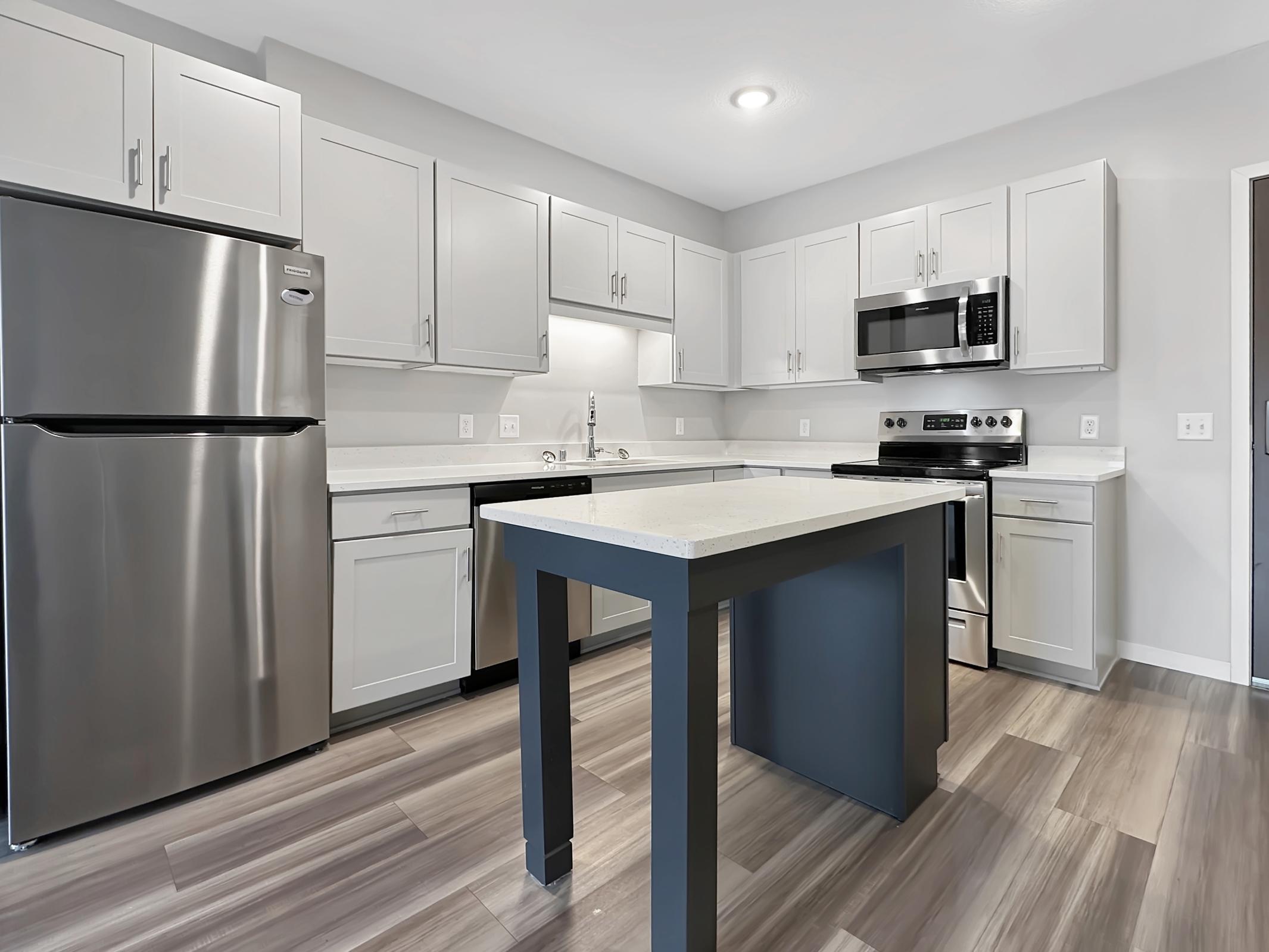 Modern kitchen featuring gray cabinets, stainless steel appliances, and a central island with a dark blue base. The layout includes a refrigerator, microwave, oven, and a sink. The flooring is wooden with a light finish, creating a bright and contemporary atmosphere.
