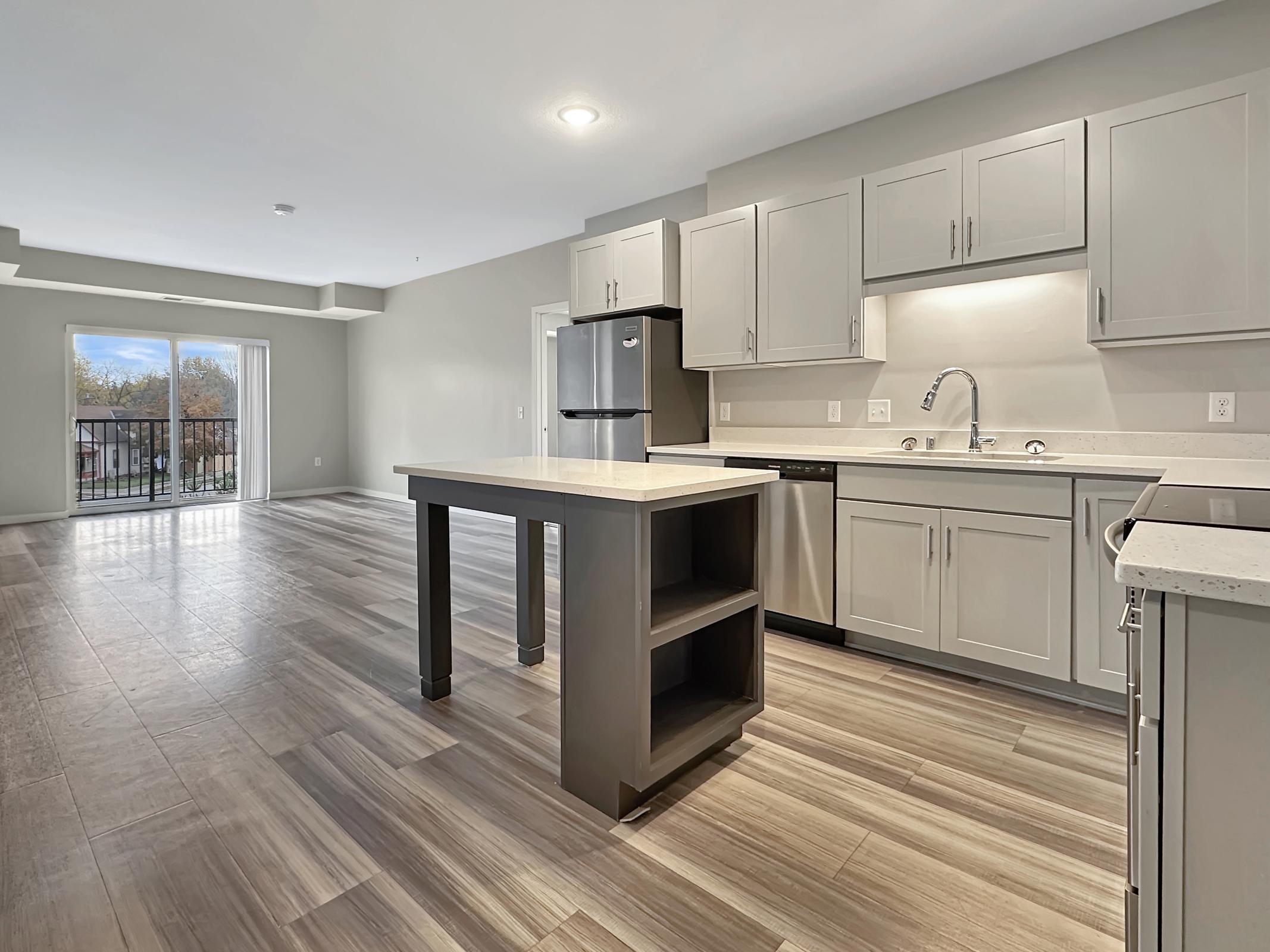Modern kitchen and living area in an apartment featuring light gray cabinets, black appliances, and a central island with a storage space. Large windows provide natural light and a view of the outdoors. The open layout enhances the spacious feel of the room.