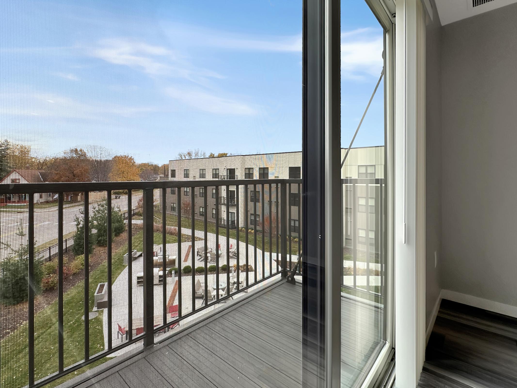 A view from a modern balcony, showcasing an outdoor courtyard with landscaped areas and seating. The sky is clear with a hint of blue, and nearby buildings feature contemporary architecture. The setting suggests a pleasant living environment with greenery and recreational space.