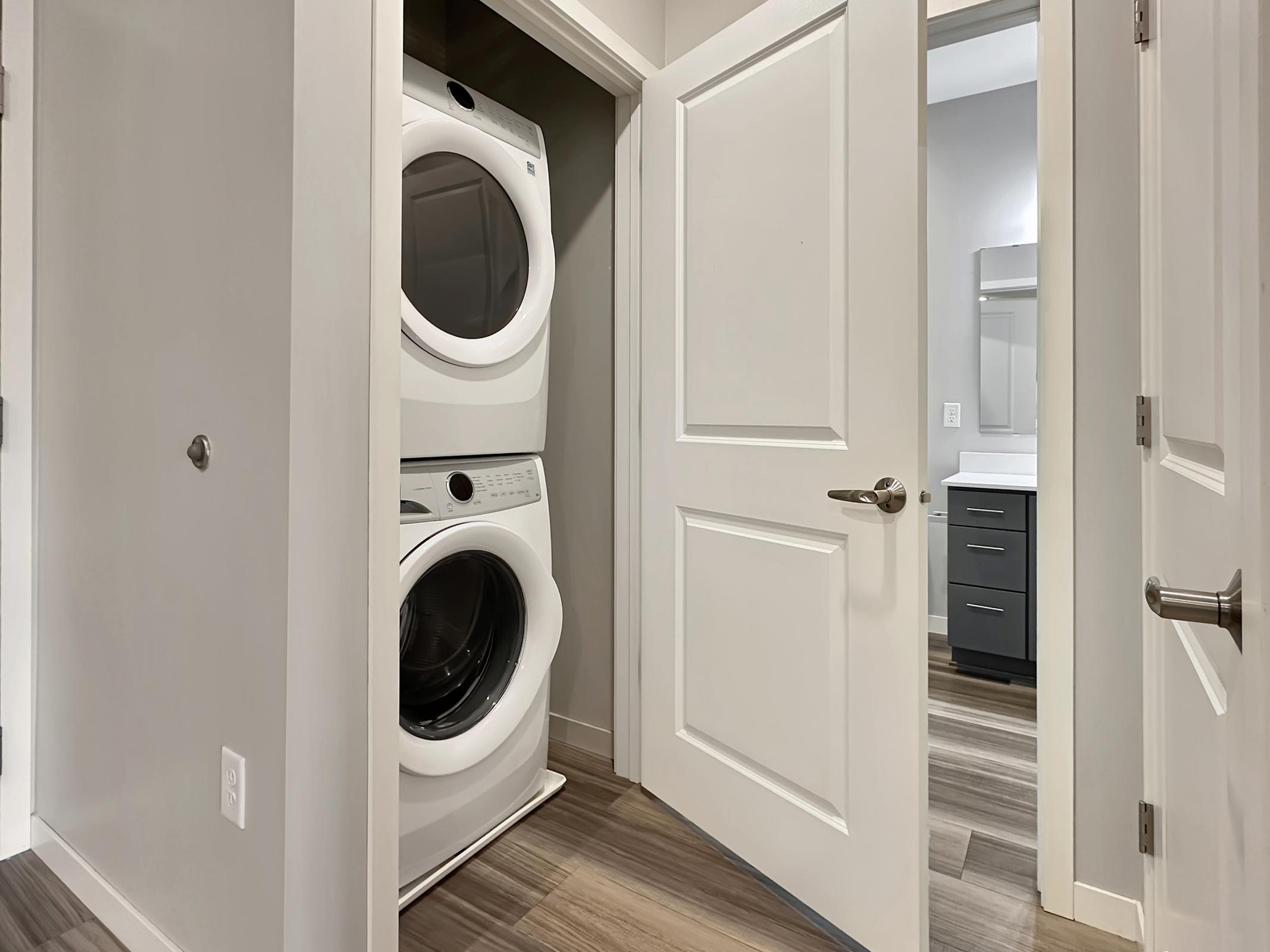 A small laundry area featuring a stacked washer and dryer, situated in a hallway with a partially open door leading to a bathroom. The walls are painted in a light neutral color, and the flooring is a wood-like laminate. There is a modern black vanity visible through the bathroom door.