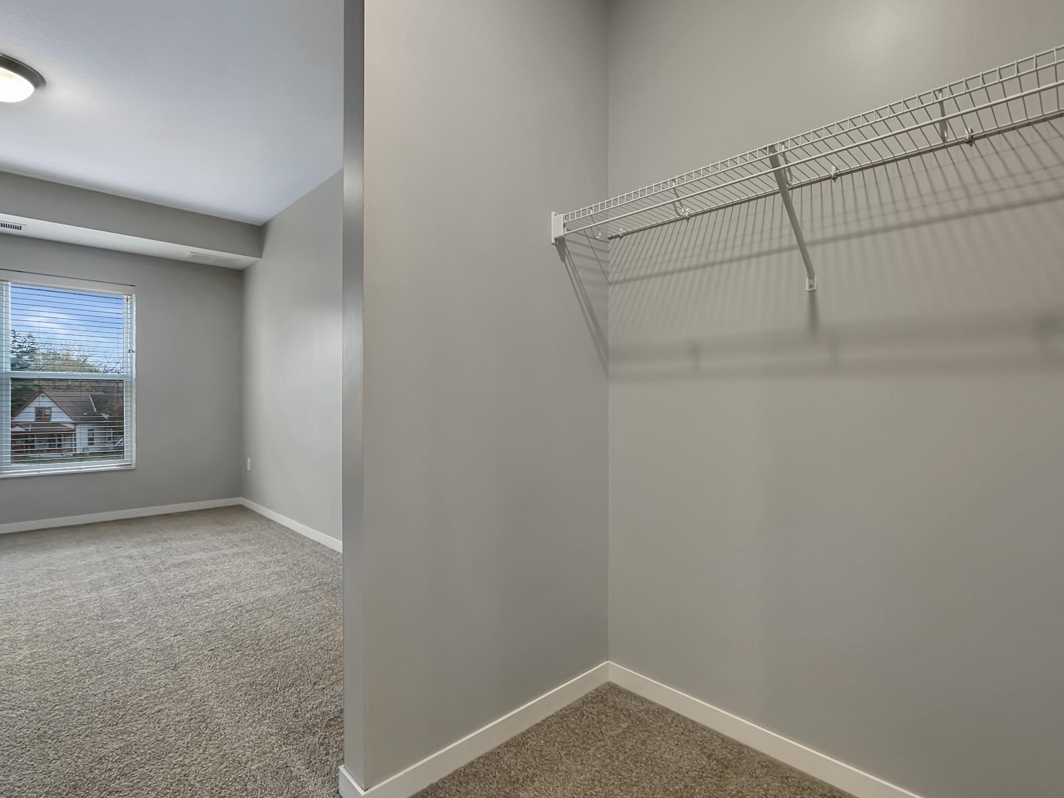 Interior view of a light gray room with a window showing houses outside. There is a carpeted area and a wire shelf mounted on the wall, indicating a closet space. The room appears freshly painted and well-lit, with a ceiling light fixture visible.