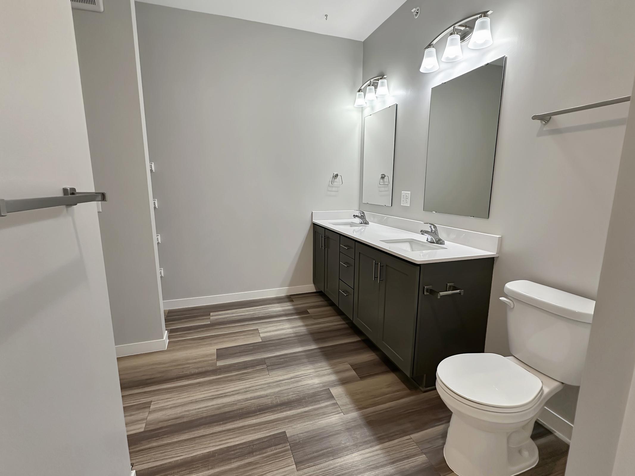 A modern bathroom featuring dual sinks with a dark wood vanity, two mirrors above the sinks, a white toilet, and stylish lighting. The floor has a wooden appearance, and the walls are painted in a light gray color, creating a sleek and contemporary look.