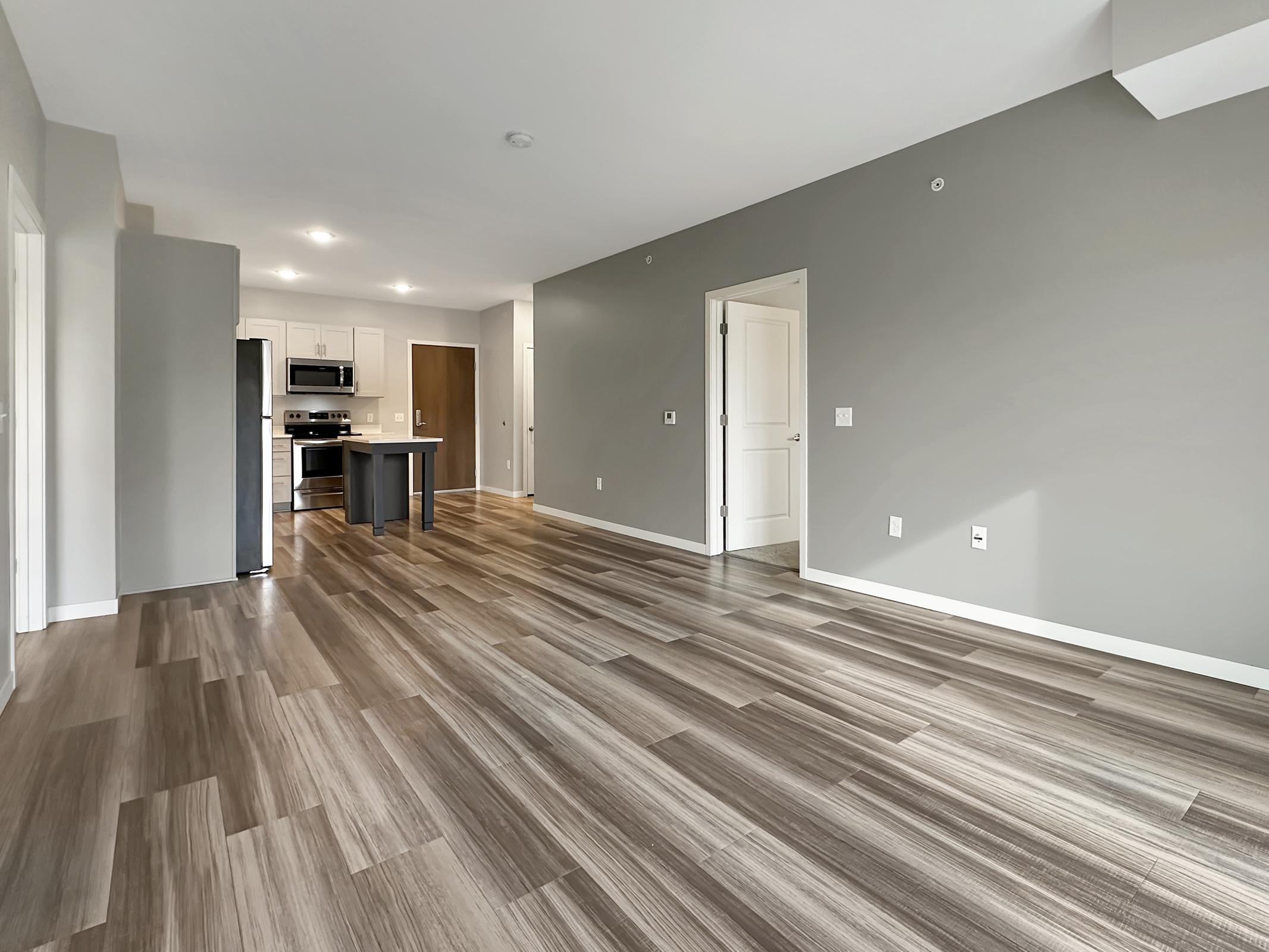 A modern, open-concept living space featuring light-colored walls and sleek wood-like flooring. The kitchen is visible in the background, equipped with dark countertops and appliances. Large windows provide natural light, enhancing the spacious feel of the room.