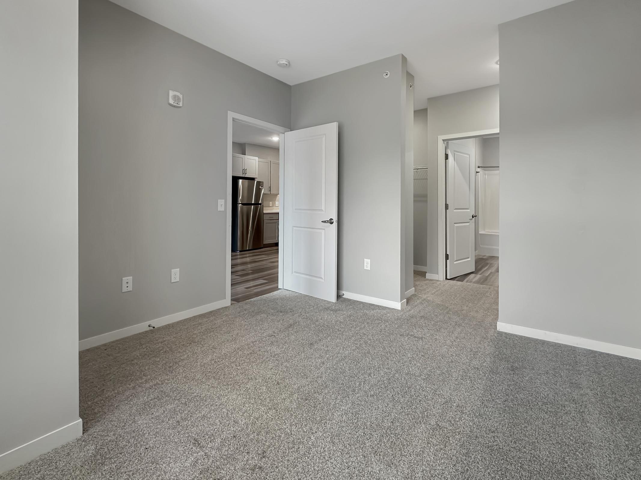 A bright, empty room with light gray walls and carpet. A doorway leads to a kitchen area visible in the background, while another doorway opens to a closet. The space features modern finishes and neutral tones, creating a welcoming atmosphere.